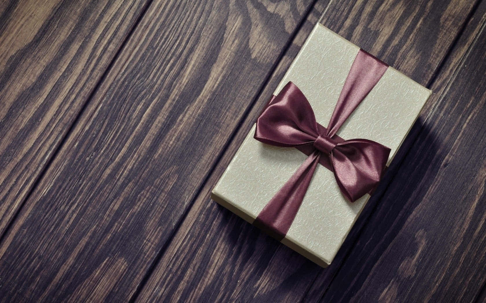 Gift 桌布 壁紙 — 一個金色禮物盒，綁著酒紅色緞帶蝴蝶結，放在深色質樸木質表面上。