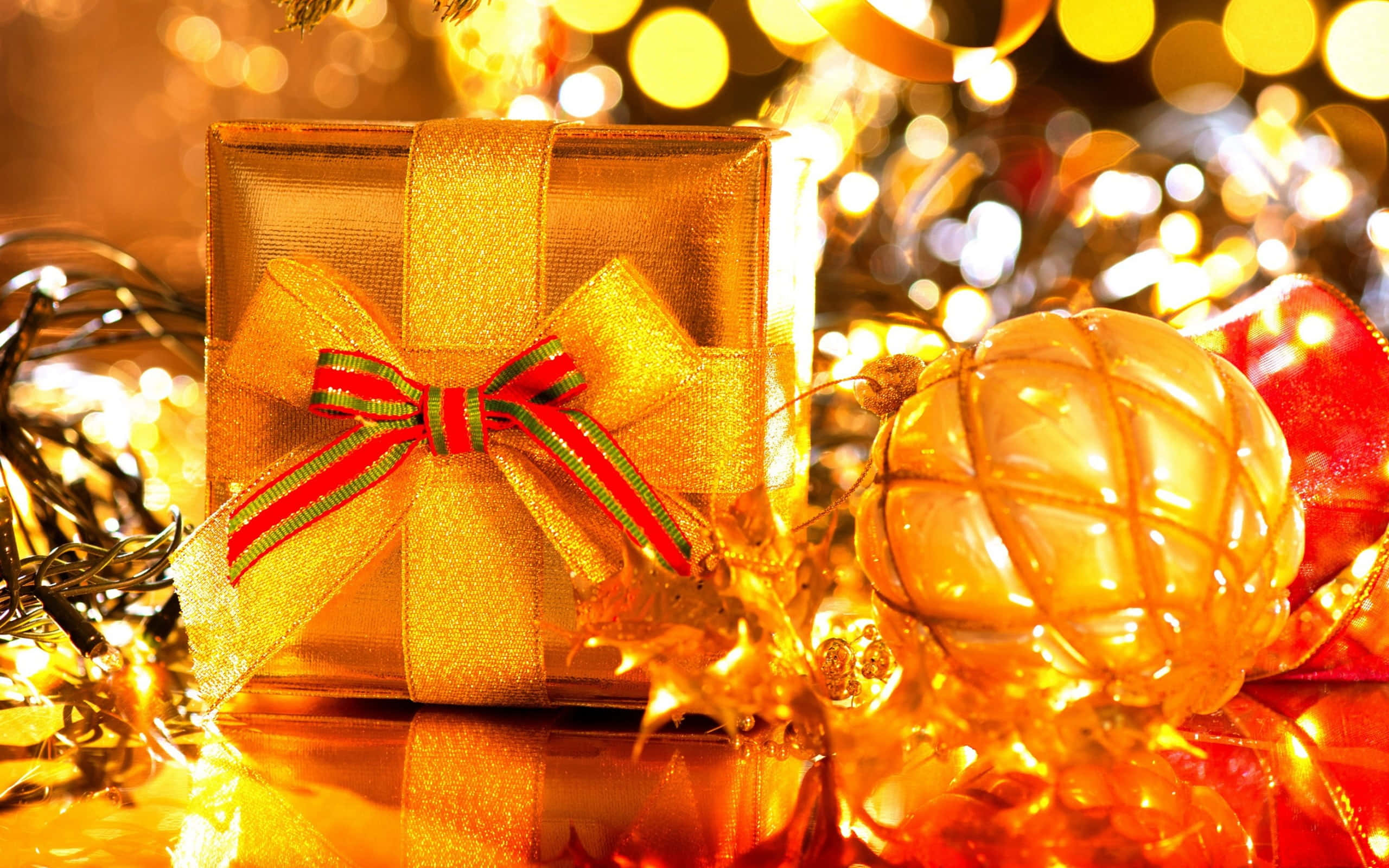 Gold Christmas 桌布 壁紙 — 一個金色禮物盒，繫著紅綠相間的蝴蝶結，放在一個閃爍的裝飾品旁邊，背景是溫暖的散景燈光。