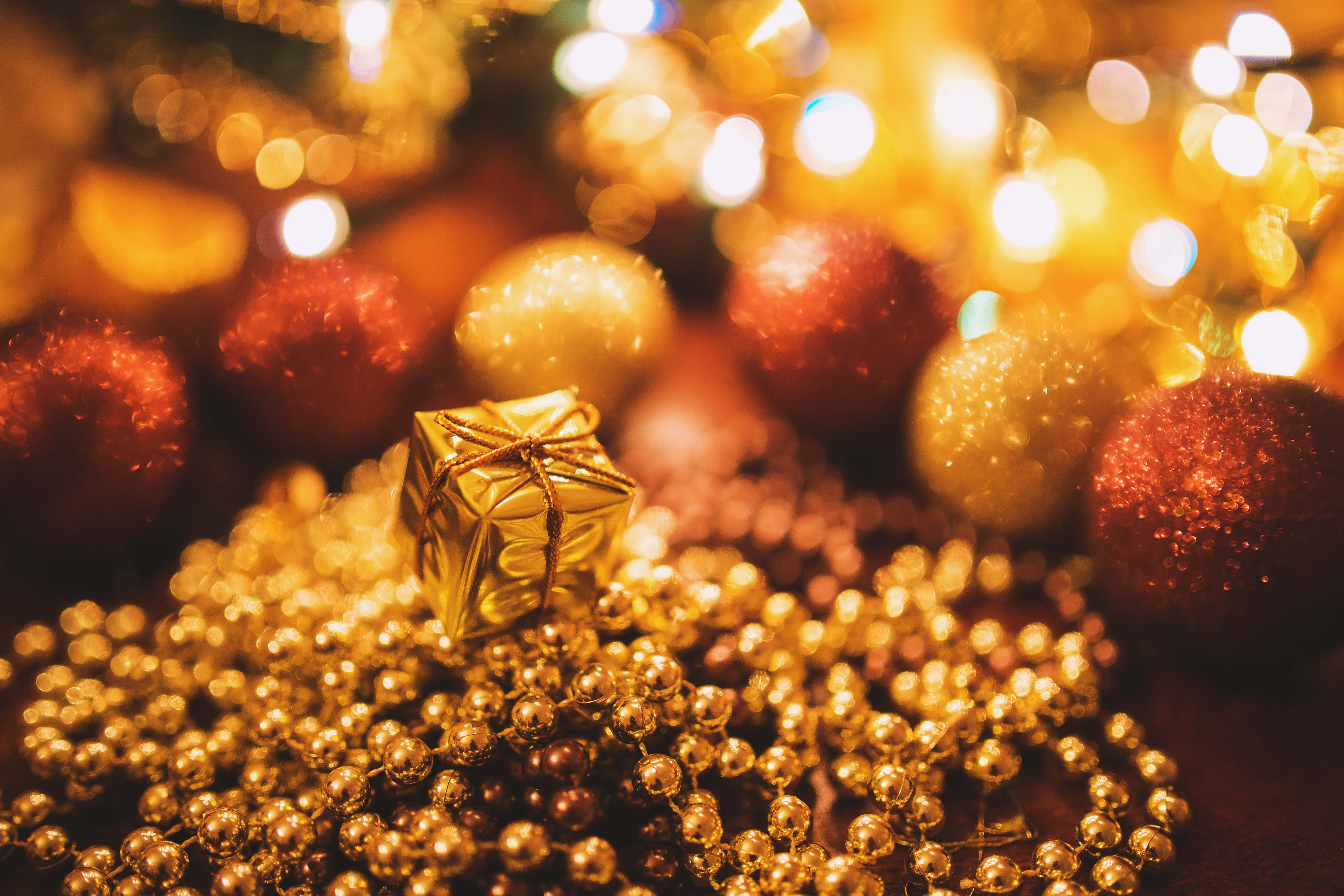 Gold Christmas 桌布 壁紙 — 一個金色禮物盒藏在閃爍的紅色和金色裝飾品和珠子中，在溫暖的散景燈光下。