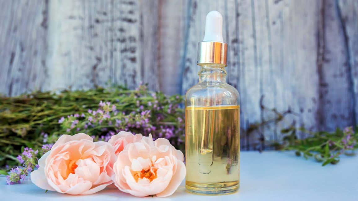 Rose Oil 桌布 壁紙 — 一個金色精油的玻璃滴管瓶，放在白色表面上，旁邊是柔和的粉紅色牡丹和紫色薰衣草，背景是風化的木頭。