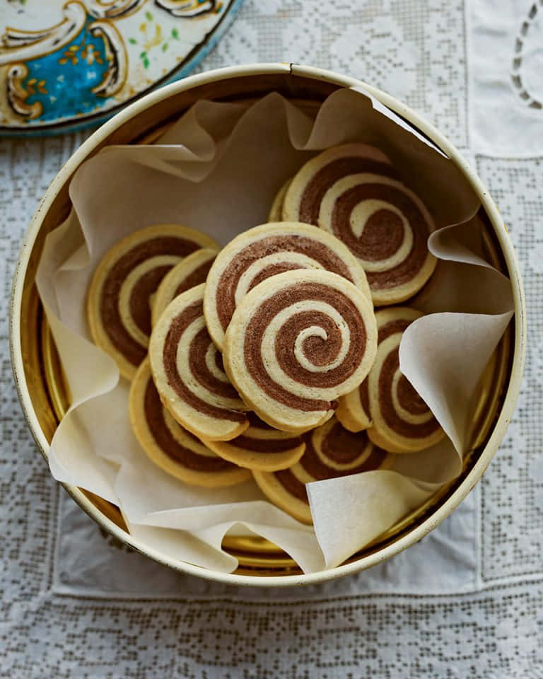 Biscuit 桌布 壁紙 — 一個金色罐子裝滿漩渦紋巧克力和香草餅乾，放在蕾絲桌布上，附近有一個裝飾盤。