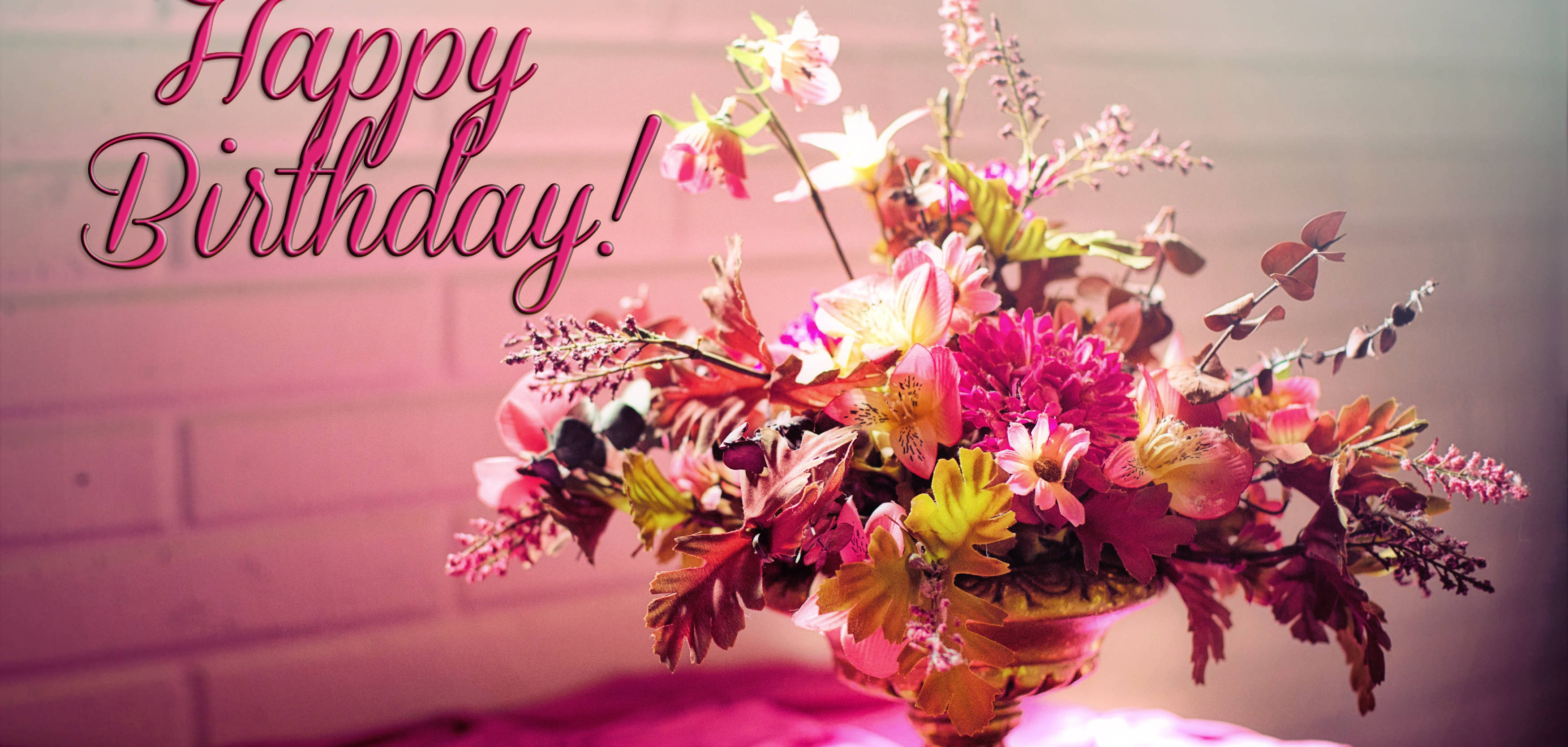 Happy Birthday Flowers 桌布 壁紙 — 一個金色花瓶裡插著粉紅色和黃色花朵的鮮豔花卉佈置，在溫暖的燈光下，以柔和的粉紅色磚牆為背景。