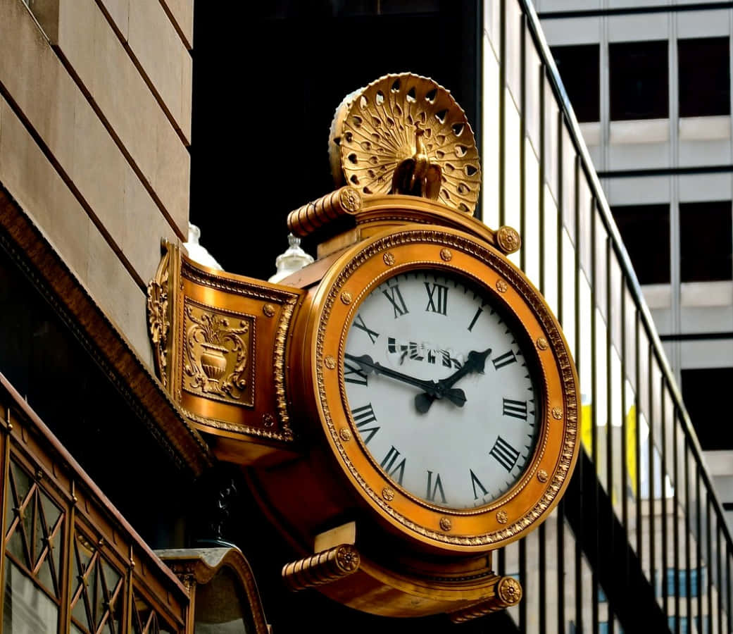 Clock 桌布 壁紙 — 一個金色華麗的時鐘，帶有孔雀頂飾，安裝在建築物外牆上，背景是現代玻璃摩天大樓。
