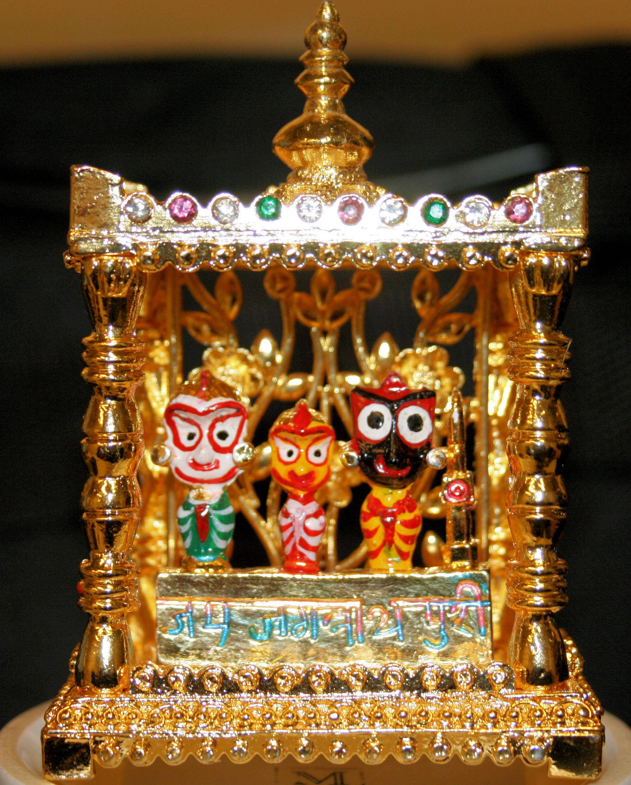 Jagannath 桌布 壁紙 — 一個金色華麗的神龕，內有三位色彩繽紛的神像，上方有珠寶裝飾的華蓋，背景深色。
