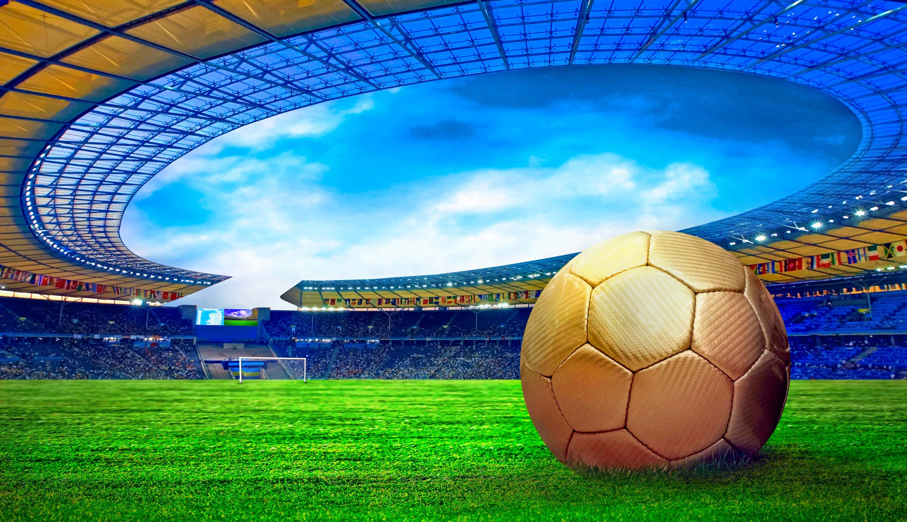 Soccer 4K 桌布 壁紙 — 一個金色足球放在一個巨大現代化體育場的鮮綠色草地上，上方是晴朗藍天，點綴著幾朵白雲。