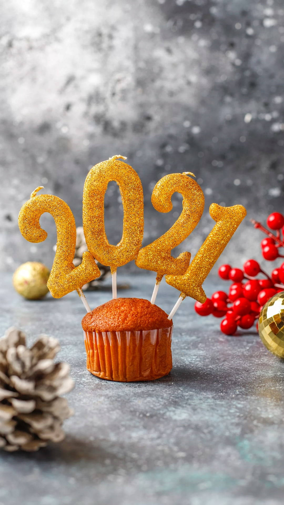 新年快樂 桌布 壁紙 — 一個金色閃亮的2021蠟燭裝飾放在一個溫暖的橙色杯子蛋糕上，周圍有紅色漿果、松果和金色飾品，置於有紋理的灰色表面上。