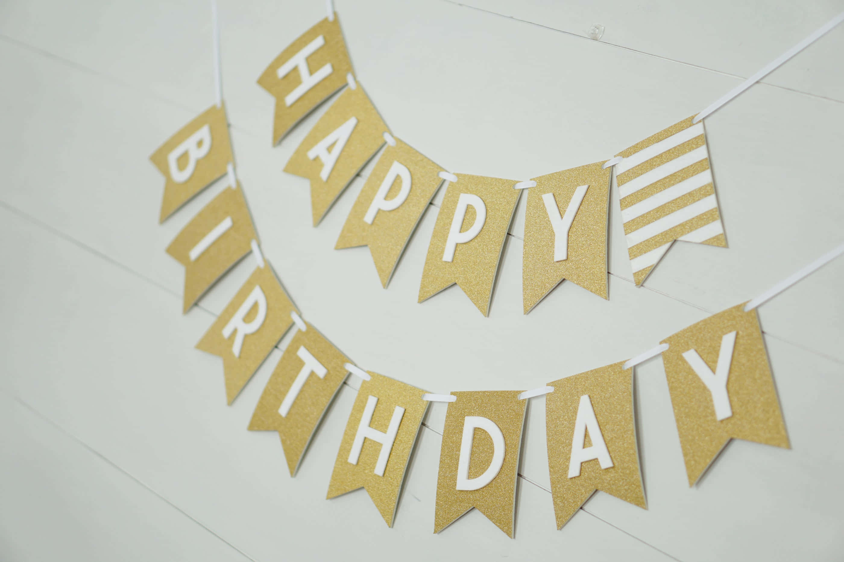 生日快樂 桌布 壁紙 — 一個金色閃亮的生日快樂橫幅，上面有白色字母，掛在淺色木牆上。