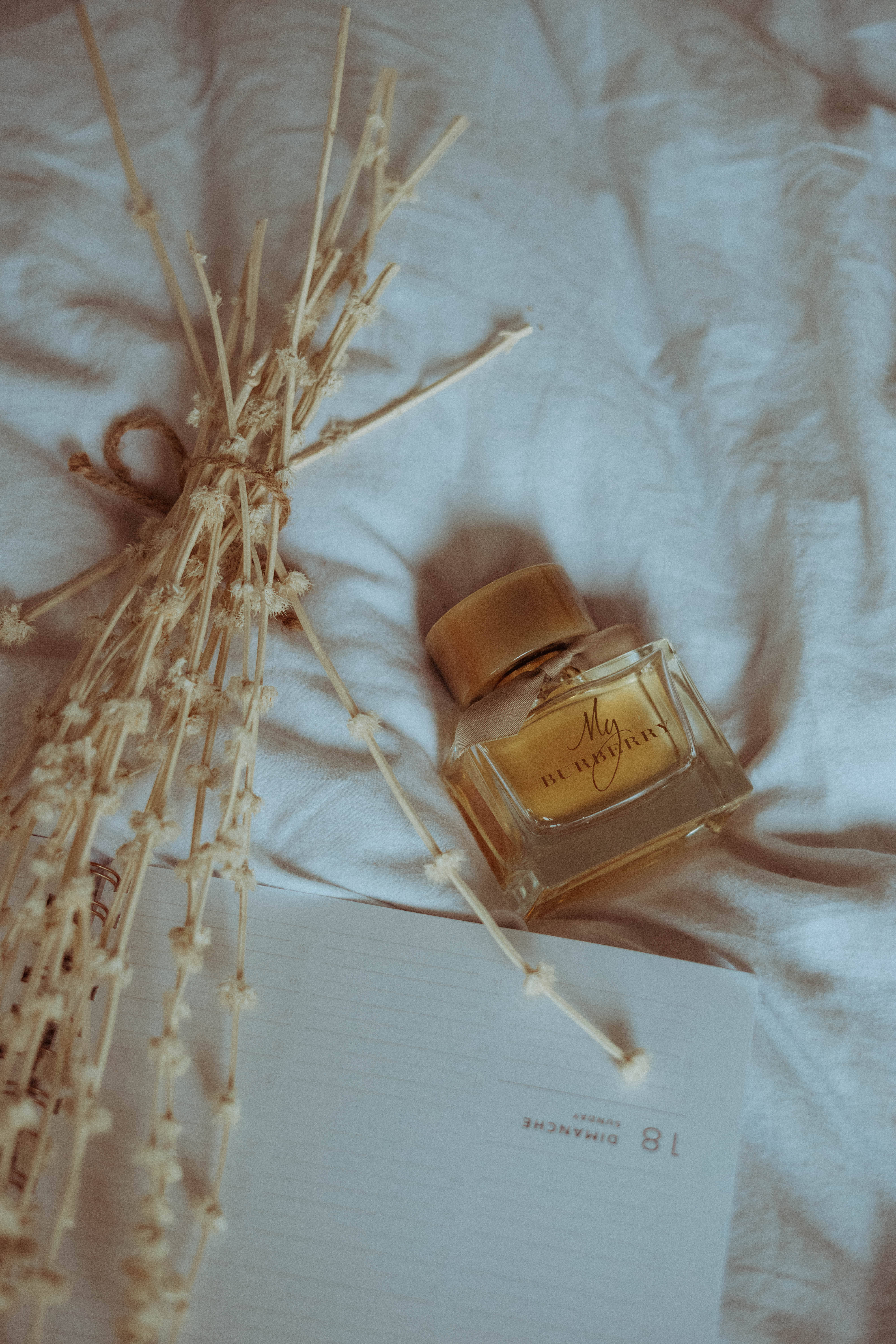 Burberry 桌布 壁紙 — 一個金色香水瓶放在乾燥薰衣草和橫線筆記本旁邊，置於柔軟皺褶的白色布料上。