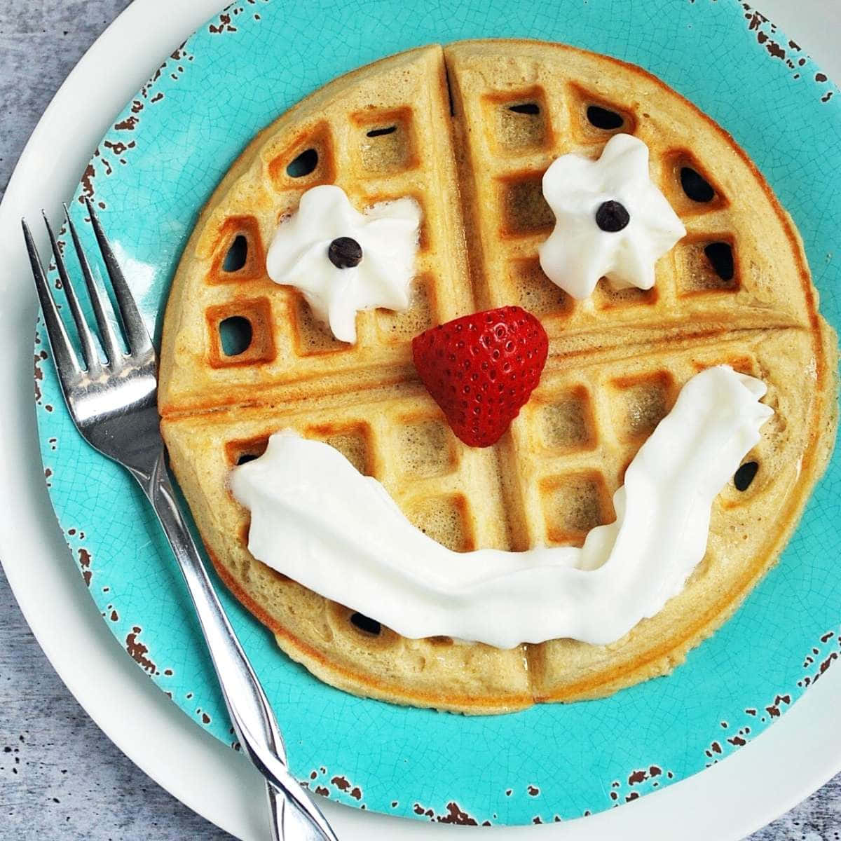 Waffle 桌布 壁紙 — 一個金色鬆餅，形狀像笑臉，有鮮奶油眼睛、草莓鼻子和寬闊的奶油笑容，放在青綠色盤子上。