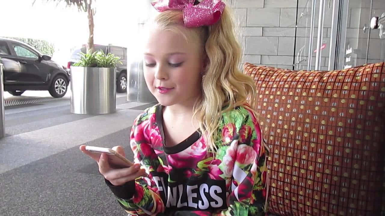 JoJo Siwa 桌布 壁紙 — 一個金髮、戴著粉紅色蝴蝶結的小女孩坐在編織長椅上，專注於手機，身處現代戶外環境。