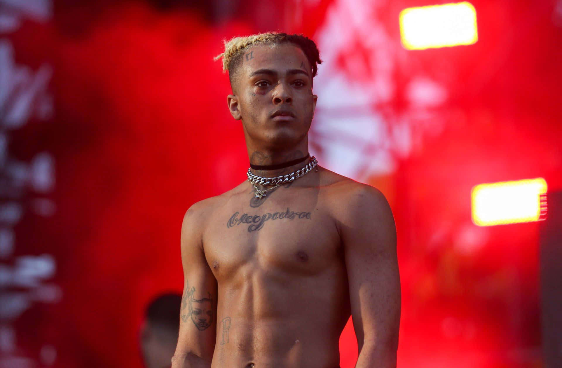 XXXTENTACION 桌布 壁紙 — 一個金髮、胸前有刺青的赤裸上身男人站在一個充滿活力的紅色舞台背景前，背景有發光的燈光。