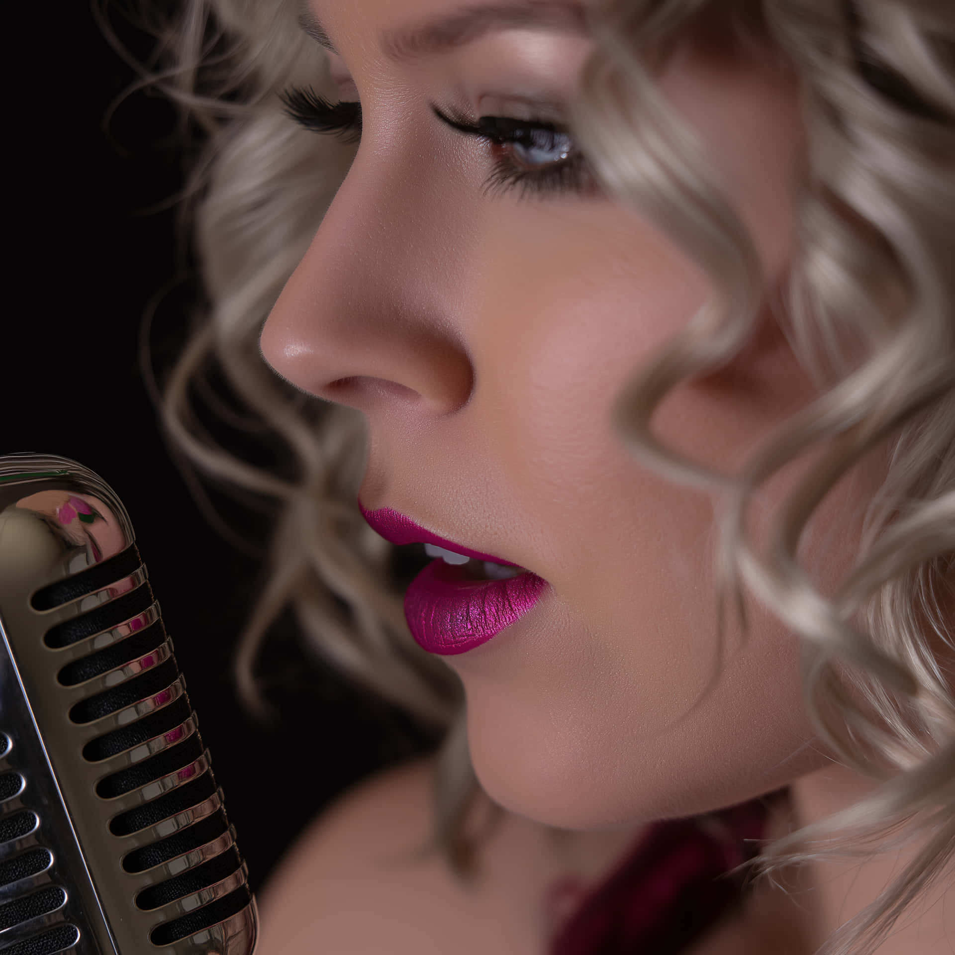 Vibrant Vocalist: Close-up of Woman Singing 壁紙 — 一個金髮女人塗著粉紅色唇膏對著復古麥克風唱歌的特寫，背景為黑色。
