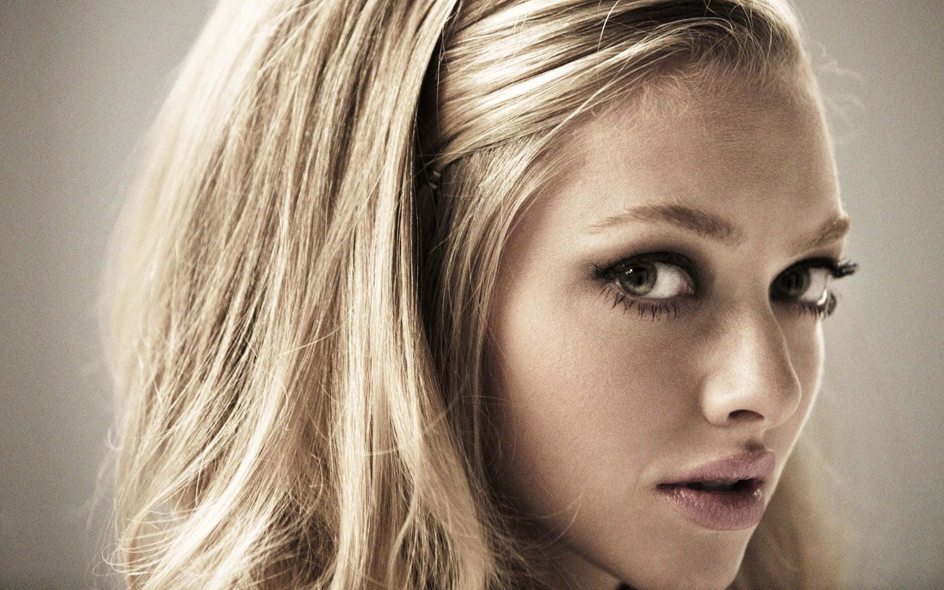 Amanda Seyfried 桌布 壁紙 — 一個金髮女性的特寫肖像，擁有強烈的綠色眼睛和柔和的光線，背景為中性色調。