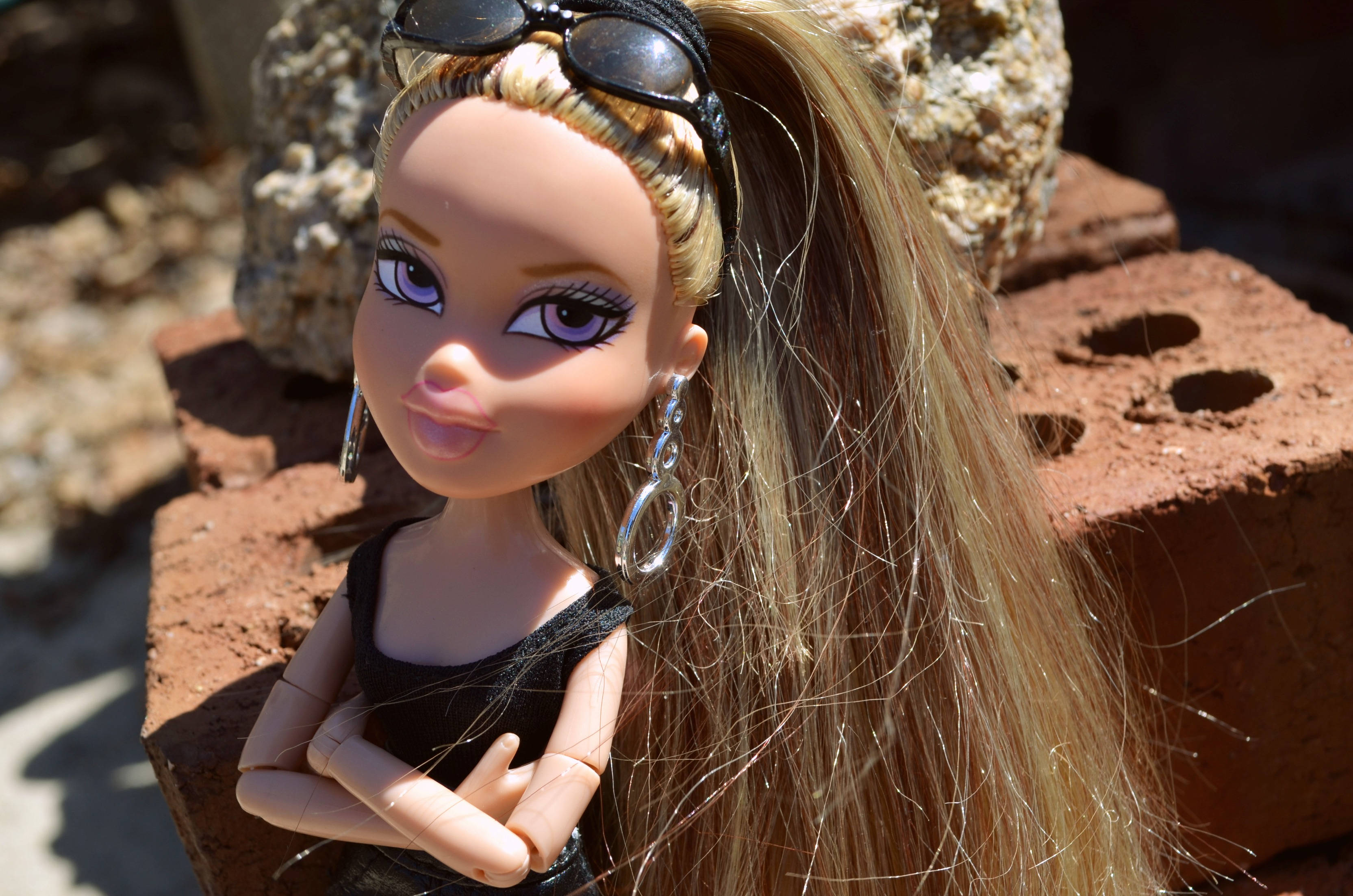 bratz 美學 桌布 壁紙 — 一個金髮娃娃頭上架著太陽眼鏡，自信地站在陽光照射的紅磚和岩石前。