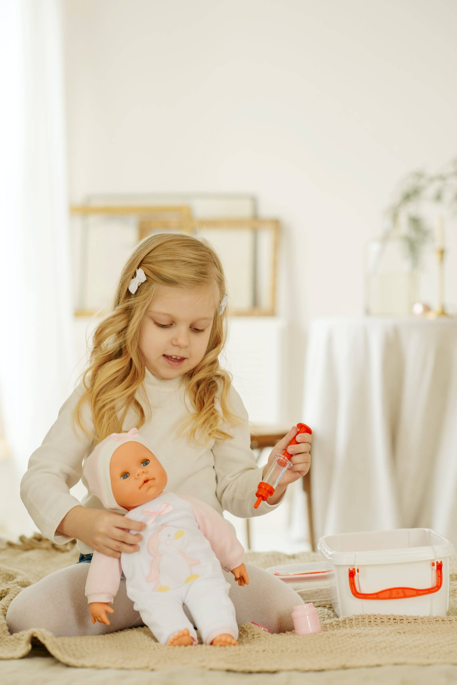Dolls 桌布 壁紙 — 一個金髮小女孩坐在地毯上，輕輕抱著一個嬰兒娃娃，同時拿著一個玩具注射器。