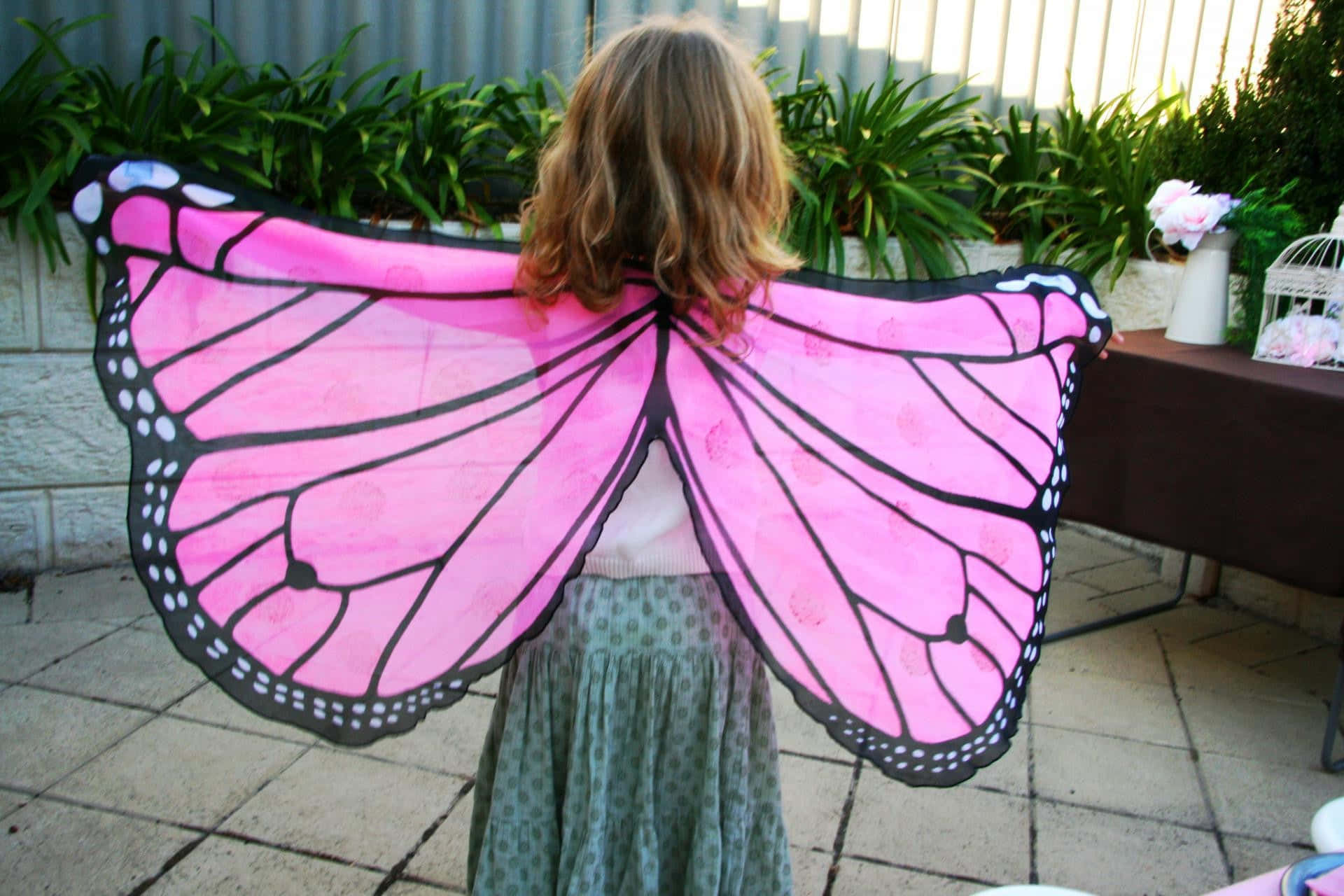 Butterfly Wing Dress 桌布 壁紙 — 一個金髮小孩戴著帶有黑色紋理和白色圓點的大型粉紅色蝴蝶翅膀，站在戶外的瓷磚庭院上。