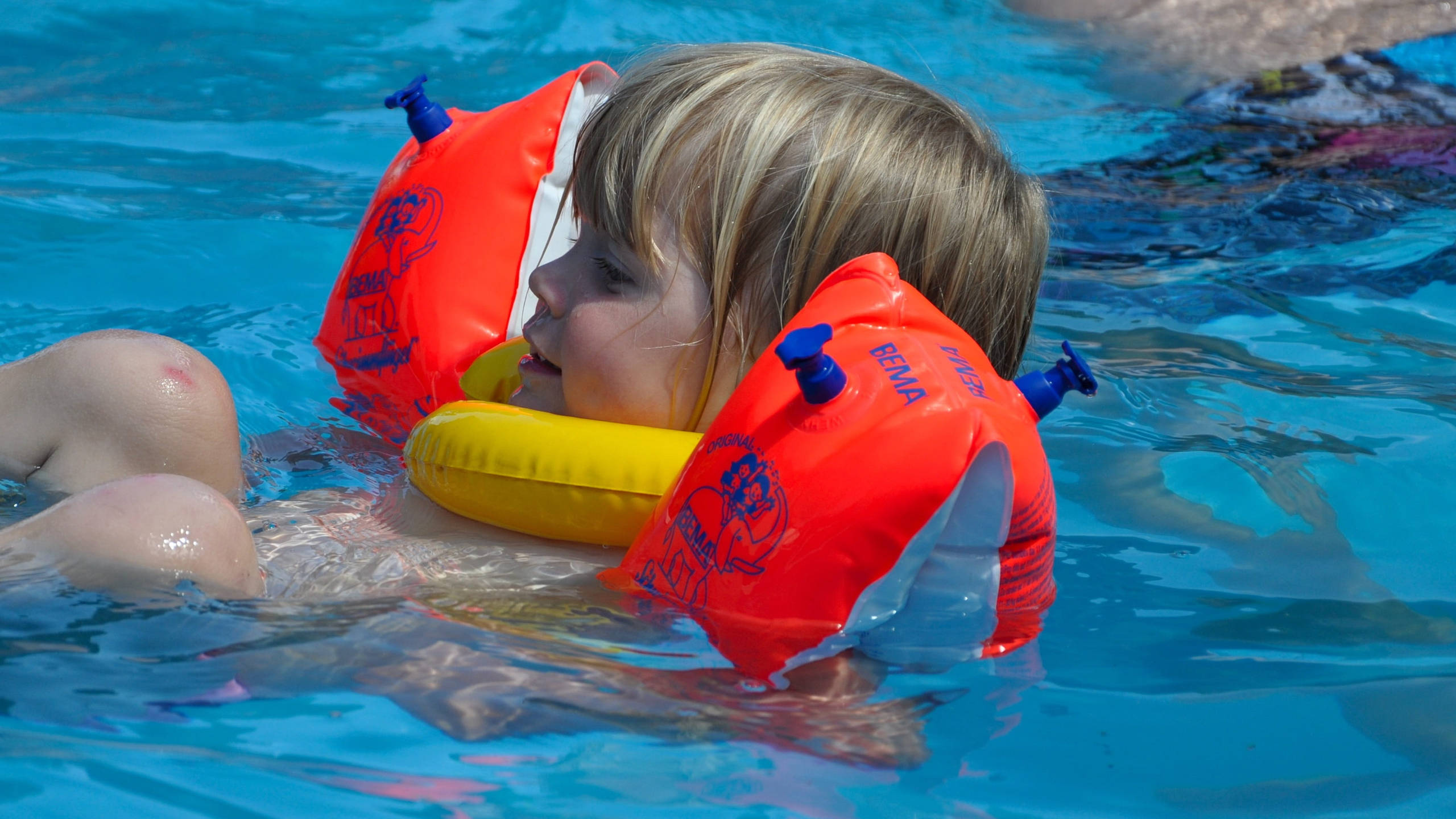 Swimming 桌布 壁紙 — 一個金髮小孩戴著橙色和黃色的充氣臂圈，在明亮的藍色泳池中快樂地漂浮。