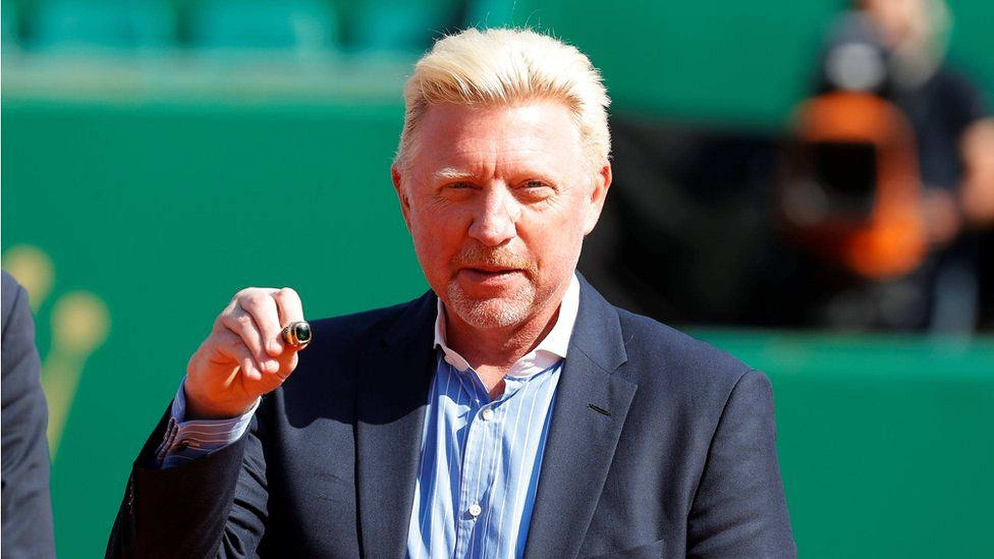 Boris Becker 桌布 壁紙 — 一個金髮男子穿著深色西裝，在戶外綠色背景下拿著筆站立。