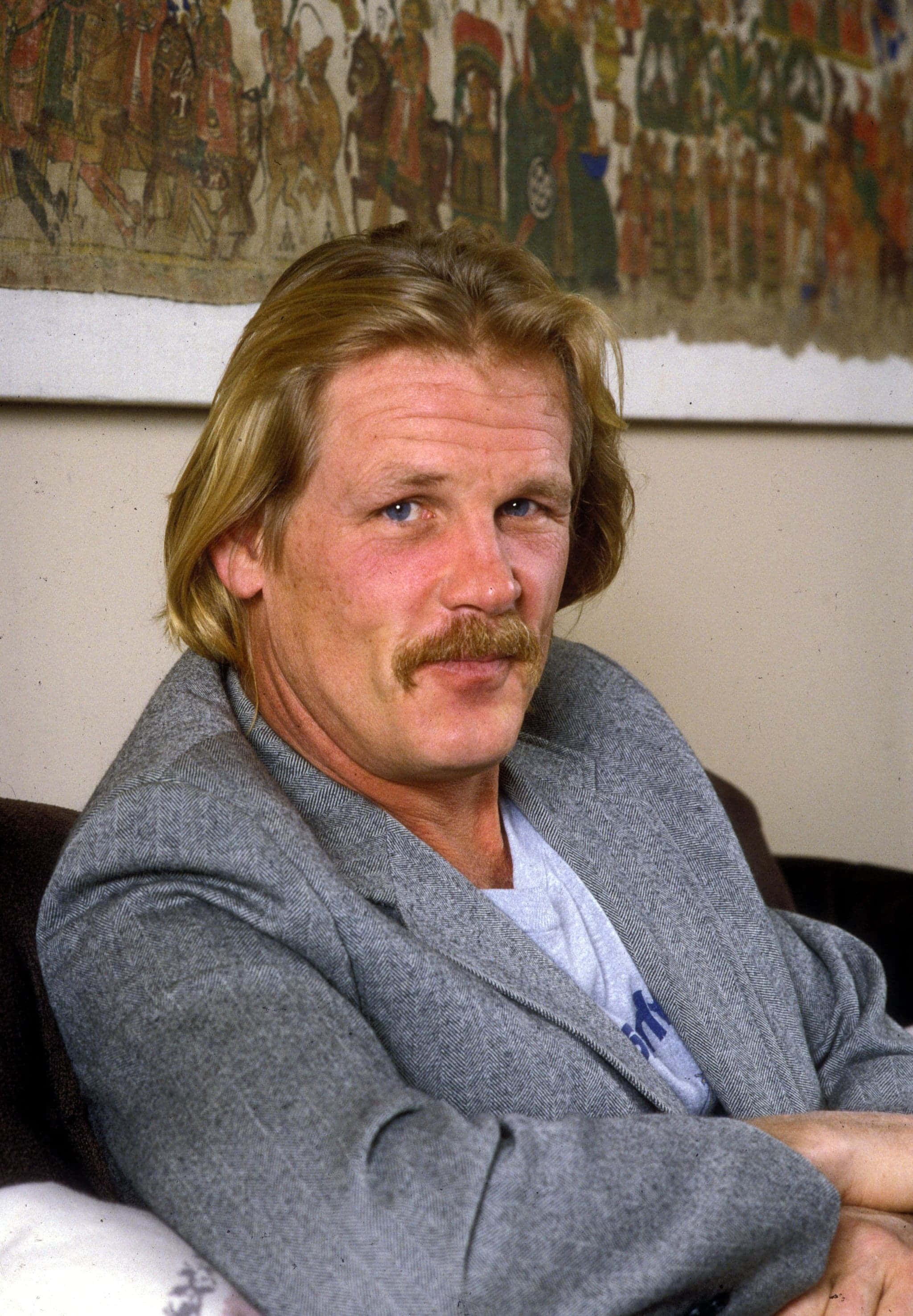 Nick Nolte 桌布 壁紙 — 一個金髮留鬍子的男人，身穿灰色西裝外套，靠著掛有彩色掛毯的牆壁，平靜地坐著。