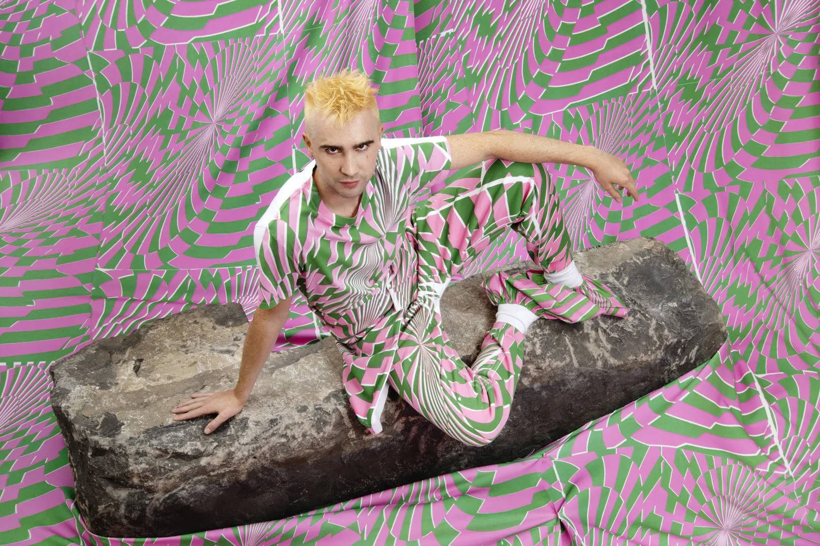Stella Mccartney 桌布 壁紙 — 一個金髮的人自信地坐在一塊大石頭上，背景是充滿活力的粉紅色和綠色幾何圖案。