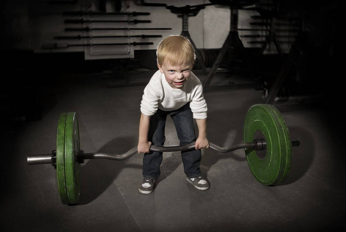 Weight Lifting 桌布 壁紙 — 一個金髮的堅定小男孩在燈光昏暗的健身房裡抓住一個帶有綠色槓片的沉重槓鈴，眼睛專注地直視前方。