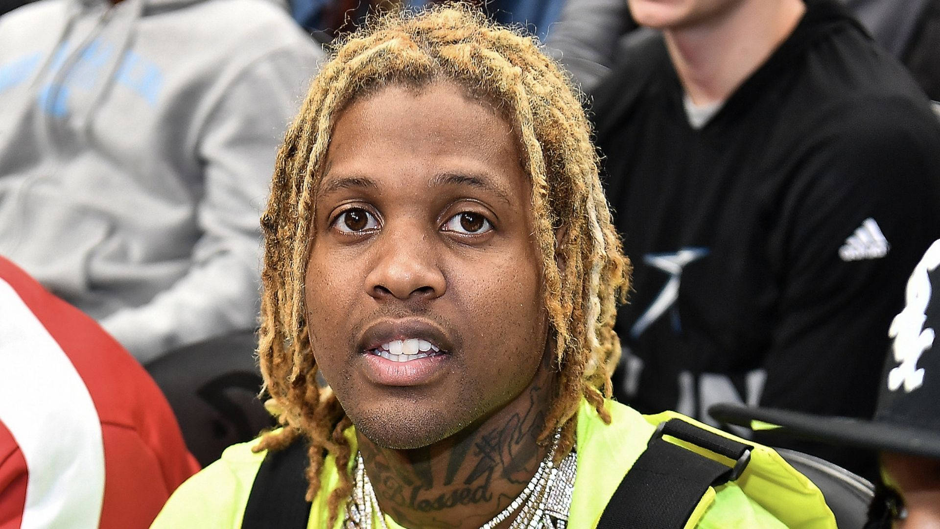 Lil Durk 桌布 壁紙 — 一個金髮辮和紋身的男子在擁擠的活動中穿著亮黃色外套的特寫。