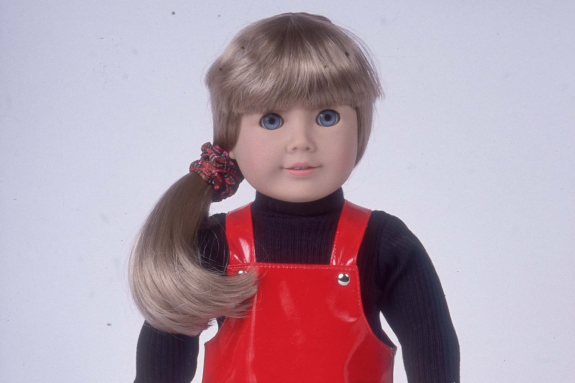 american girl 桌布 壁紙 — 一個金髮馬尾辮藍眼睛的復古娃娃，在純白背景前穿著黑色高領衫外搭閃亮紅色連身褲。