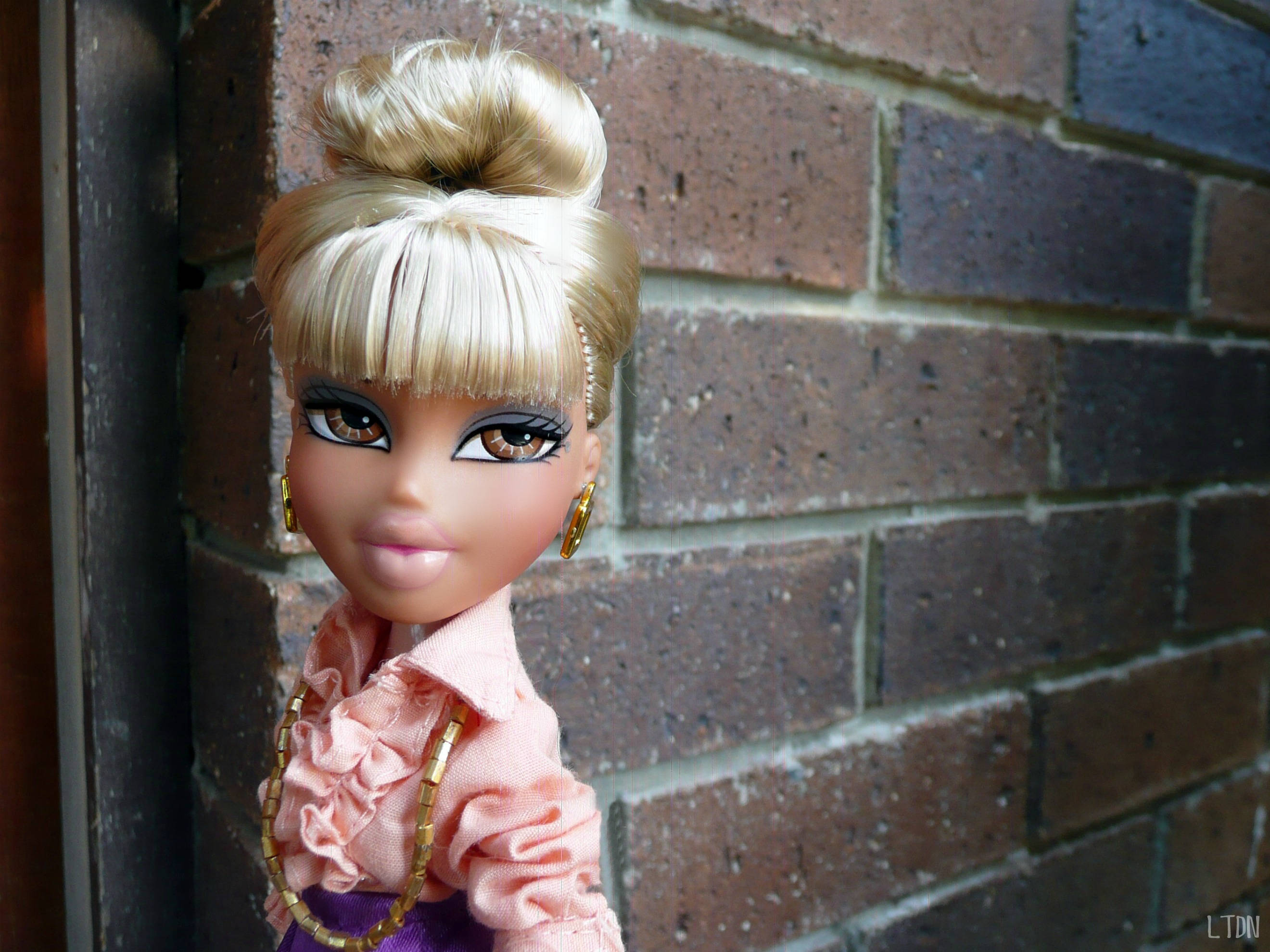 bratz 美學 桌布 壁紙 — 一個金髮高髻和瀏海的娃娃，站在有紋理的磚牆前，穿著粉紅色荷葉邊上衣和金色珠寶。