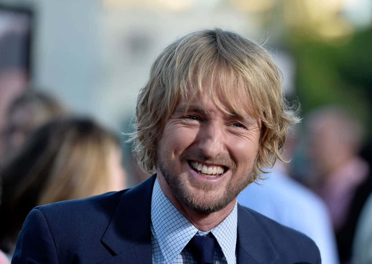 Owen Wilson 桌布 壁紙 — 一個金髮鬍鬚的男人在模糊的人群背景下，穿著深色西裝打領帶，露出燦爛的笑容。
