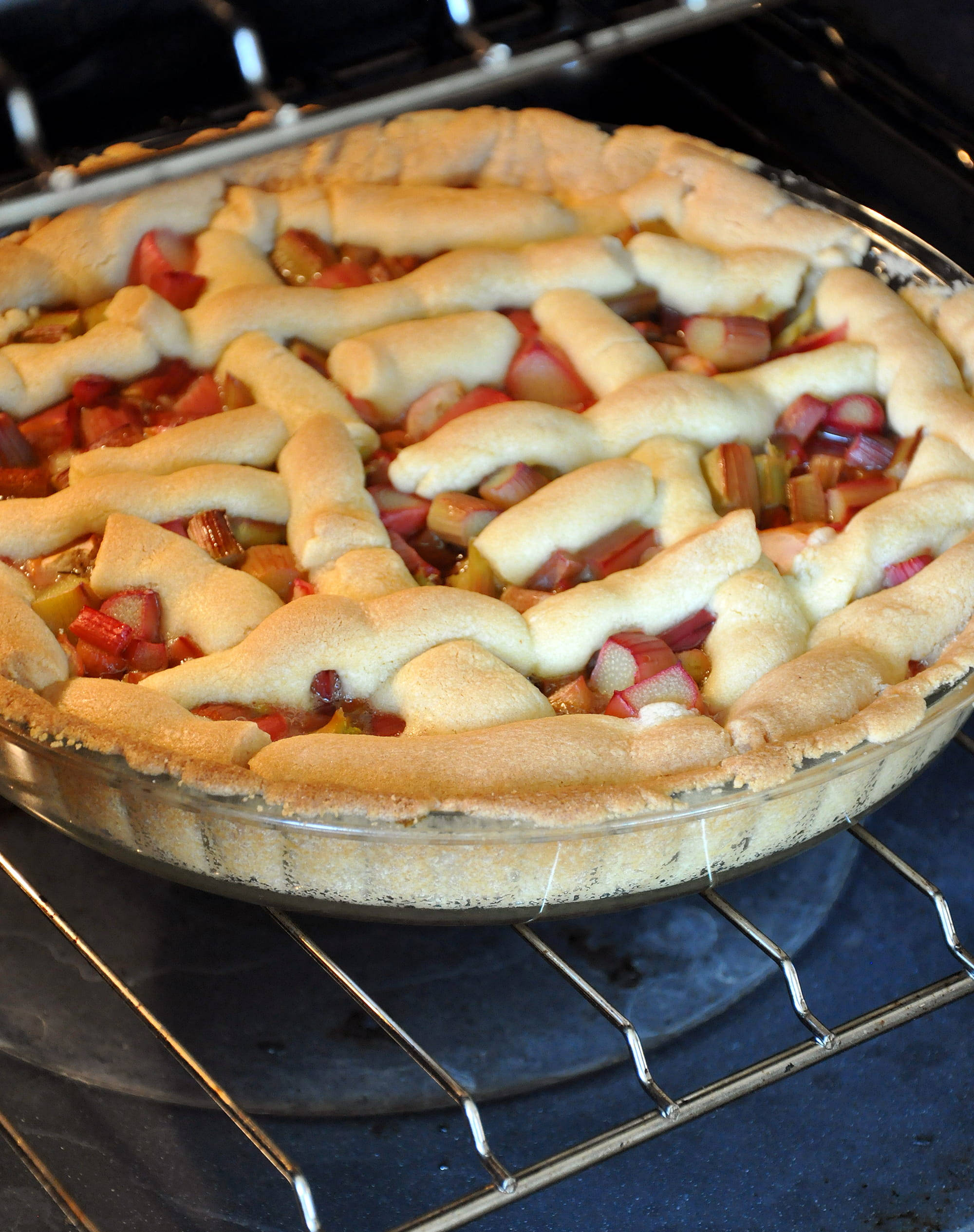 Pie 桌布 壁紙 — 一個金黃色、頂部有格子狀的烤大黃派在玻璃盤中，放在烤箱架上烘烤，蒸氣上升。