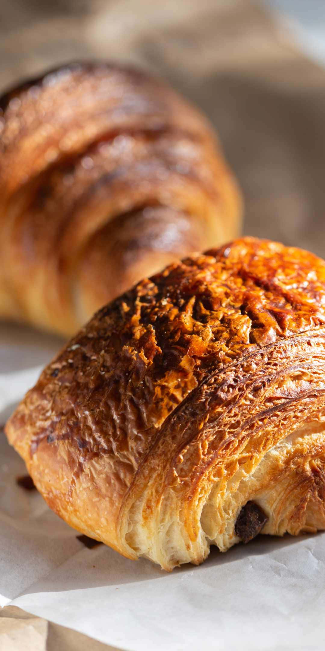 Pastry 桌布 壁紙 — 一個金黃色可頌的近距離特寫，帶有酥脆的層次和焦糖糖衣，放在烘焙紙上。