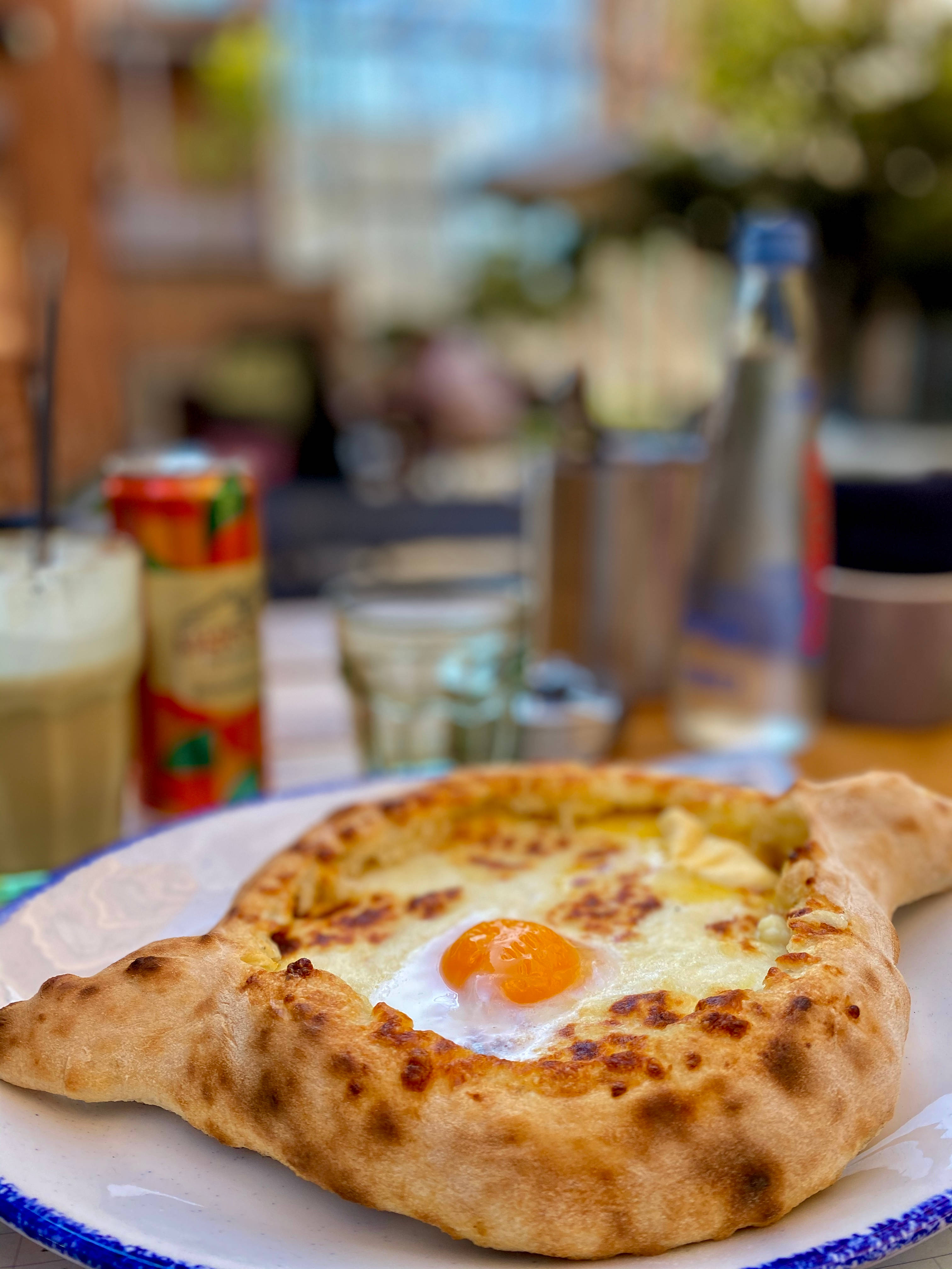 Cheese 桌布 壁紙 — 一個金黃色的 khachapuri 配上流心蛋黃，放在藍邊白盤上，周圍有飲料和調味品，在明亮的戶外環境中。