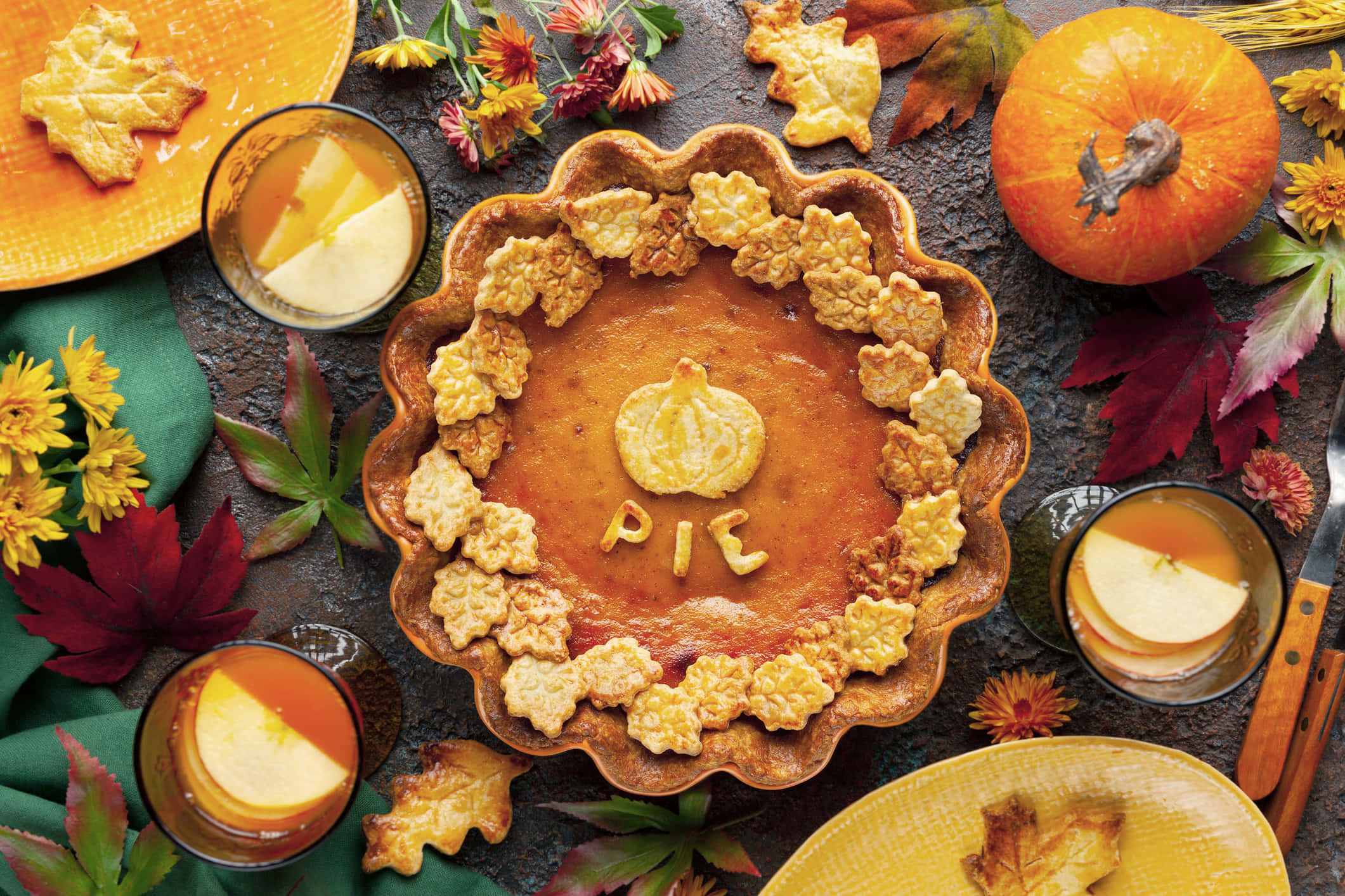 Fall Thanksgiving 桌布 壁紙 — 一個金黃色的南瓜派，上面有葉形餅乾和PIE字樣，周圍環繞著秋葉、南瓜和蘋果酒杯子。