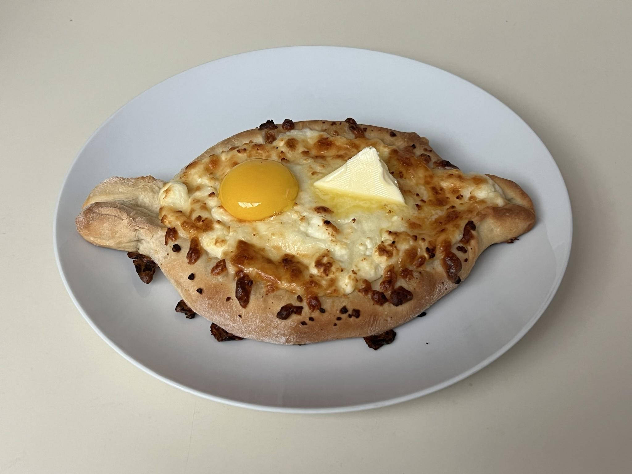 Cheese 桌布 壁紙 — 一個金黃色的哈恰普里麵包，上面有融化起司、一個亮黃色的蛋黃和一小塊奶油，放在白色盤子上。