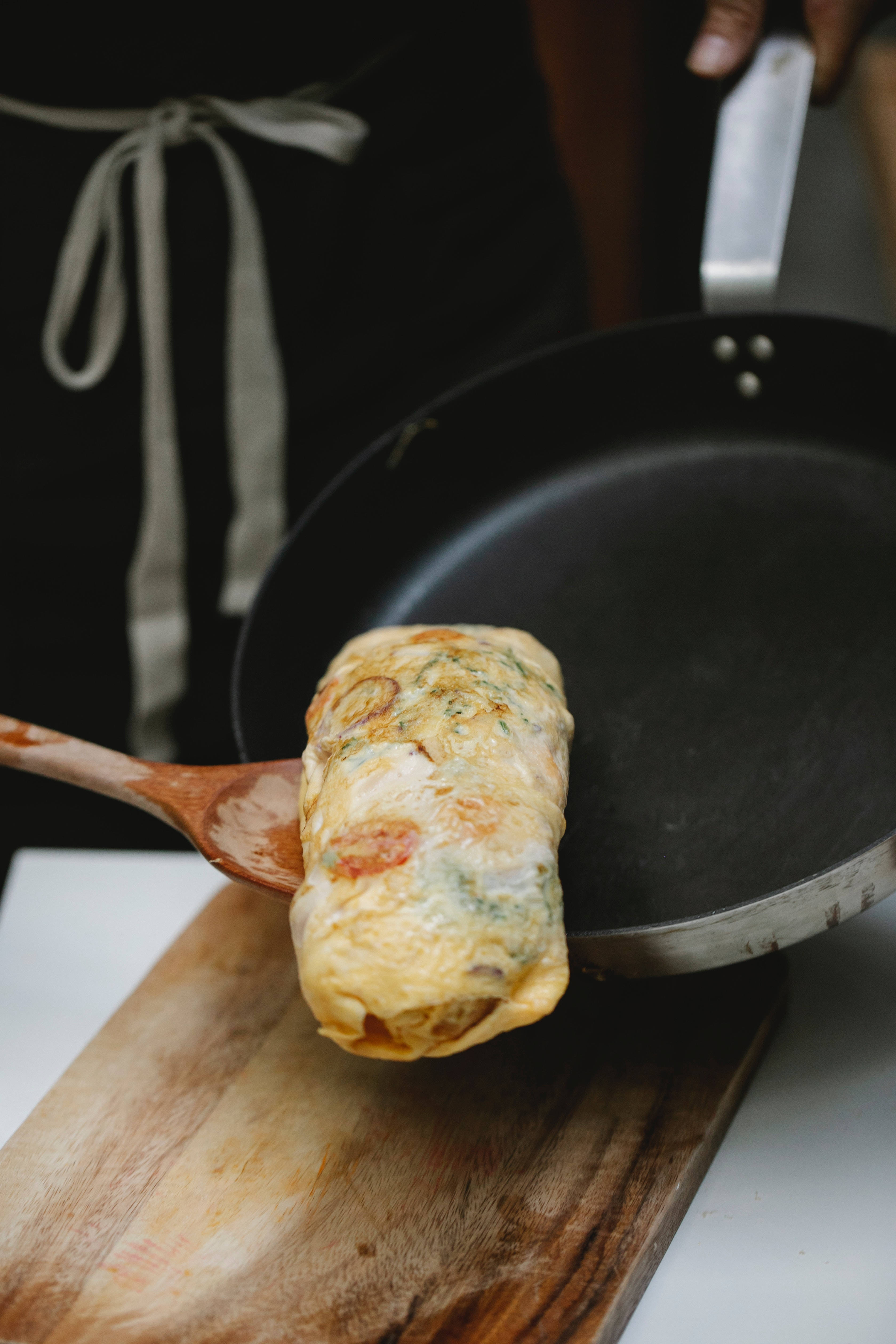 Egg 桌布 壁紙 — 一個金黃色的歐姆蛋從黑色煎鍋中用木鍋鏟捲起並提起，放在一個質樸的木板上。