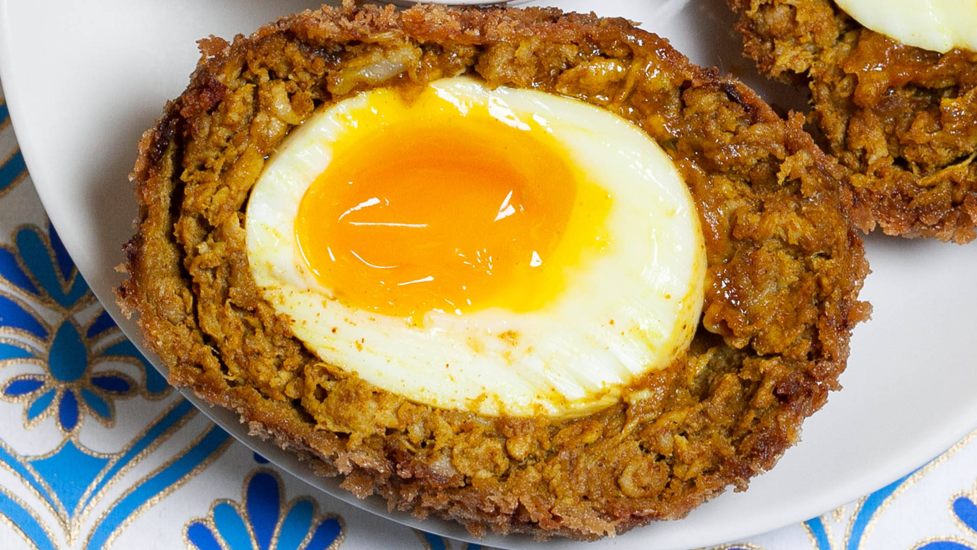 Egg 桌布 壁紙 — 一個金黃色的流心蛋黃位於一個酥脆金黃色蘇格蘭蛋的中心，放在一個有藍色圖案布的白色盤子上。