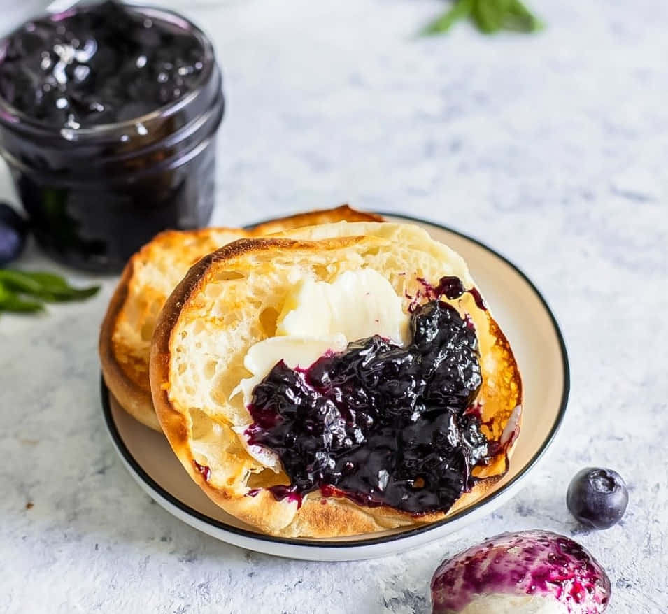 Blueberry Jam 桌布 壁紙 — 一個金黃色的烤麵包卷切開，配上奶油狀的牛油和濃郁的深色藍莓果醬，放在一個有斑點的盤子上。
