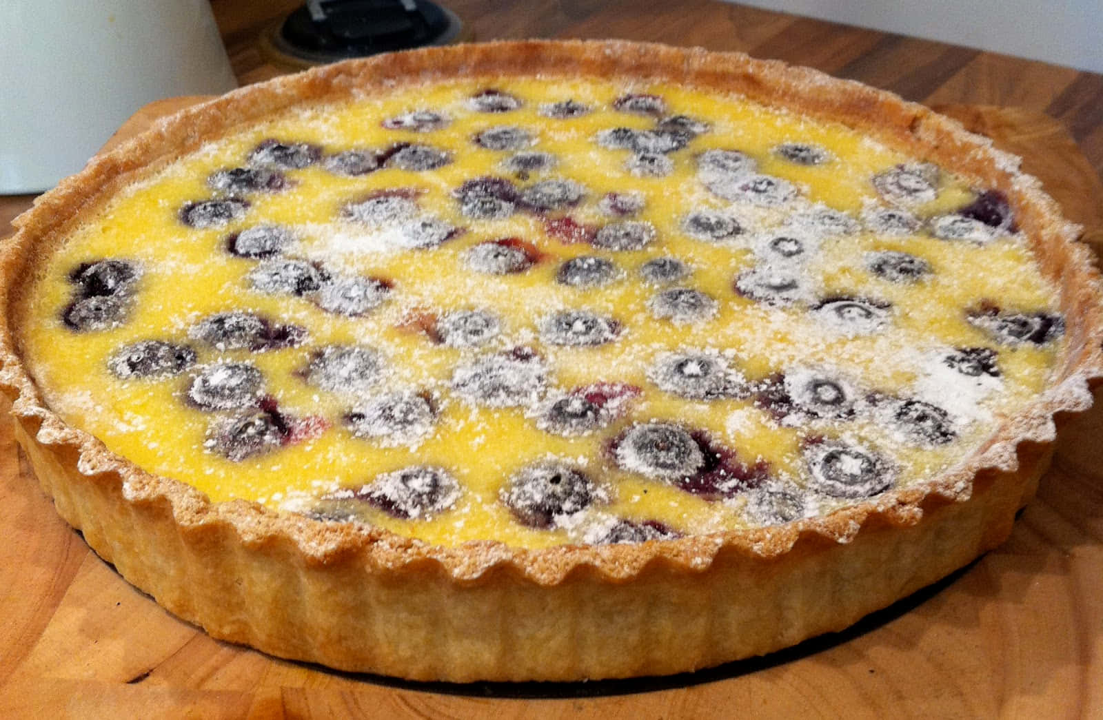 Blueberries Tart 桌布 壁紙 — 一個金黃色的藍莓塔撒上糖粉，放在木製砧板上。