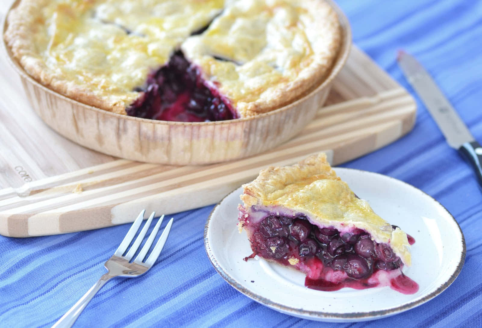 Blueberry Pie 桌布 壁紙 — 一個金黃色的藍莓派，帶有酥脆的外皮，放在木盤中，旁邊是一塊放在盤子上的切片，置於藍色條紋布上。