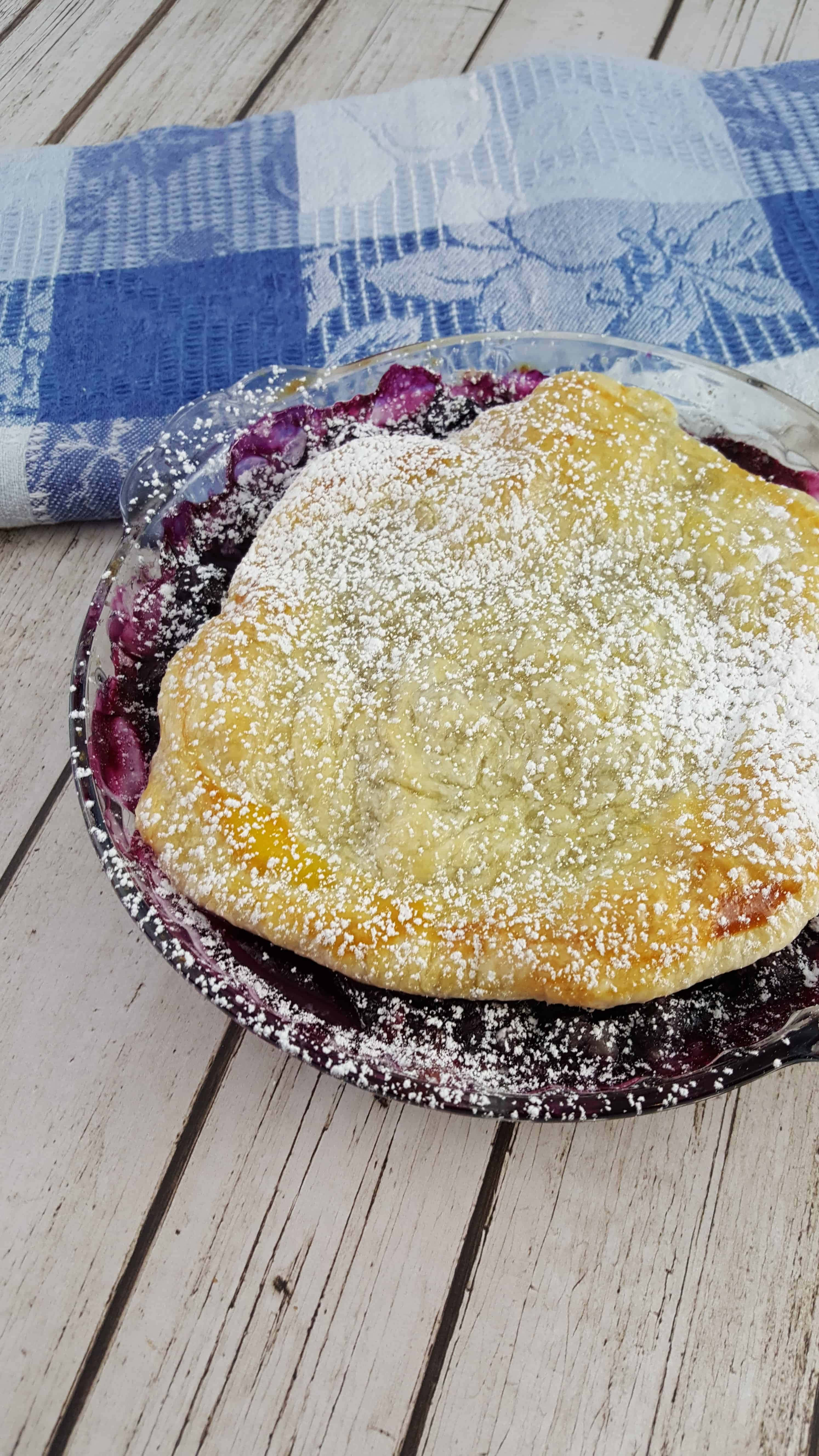 Blueberries Tart 桌布 壁紙 — 一個金黃色的藍莓派撒上糖粉，放在玻璃盤中，置於一張風化的木桌上，旁邊有一塊藍色格子布。