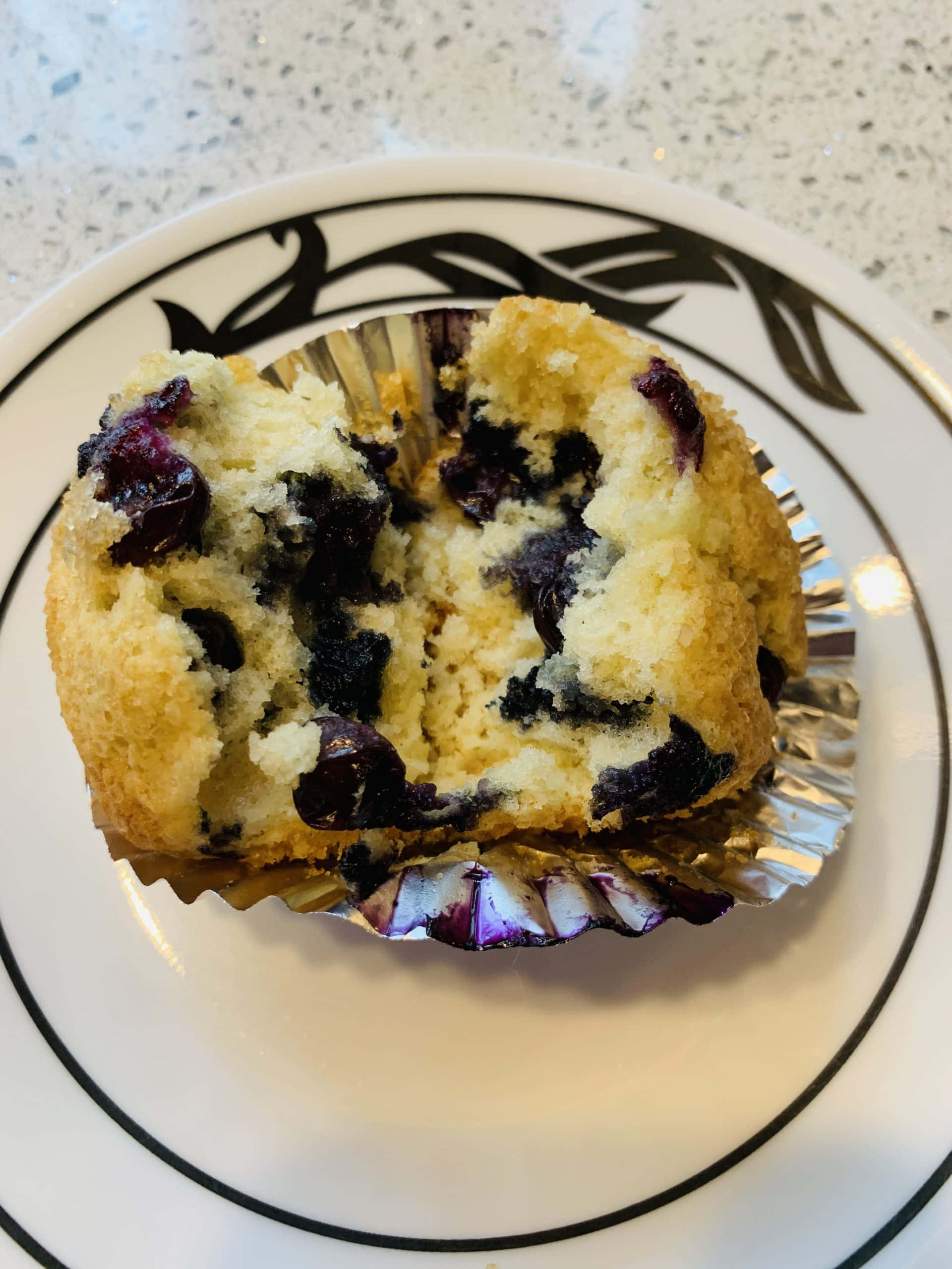 Blueberry Muffins 桌布 壁紙 — 一個金黃色的藍莓馬芬被撕開，露出多汁的莓果和柔軟的碎屑，放在有圖案的盤子上。