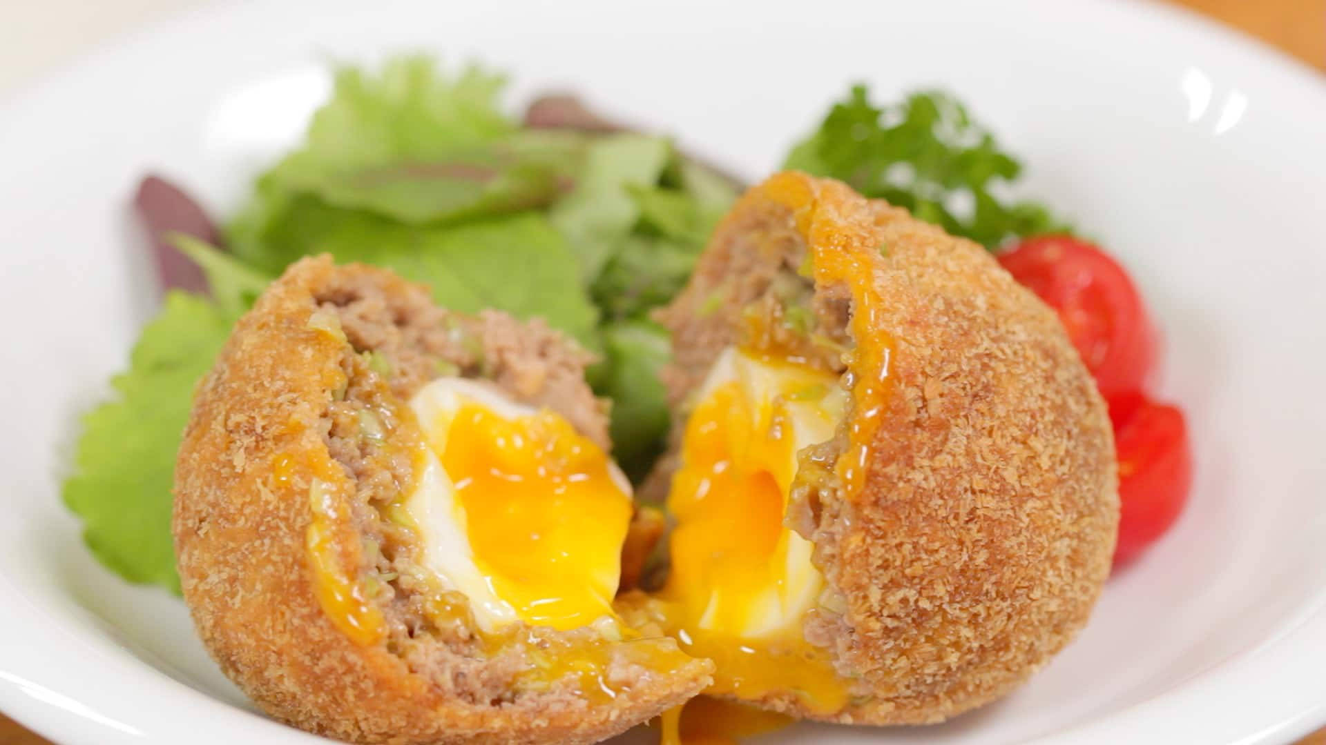 Egg 桌布 壁紙 — 一個金黃色的蘇格蘭蛋切開，露出流心蛋黃藏在美味的肉中，配上新鮮蔬菜和櫻桃番茄，放在白色盤子上。