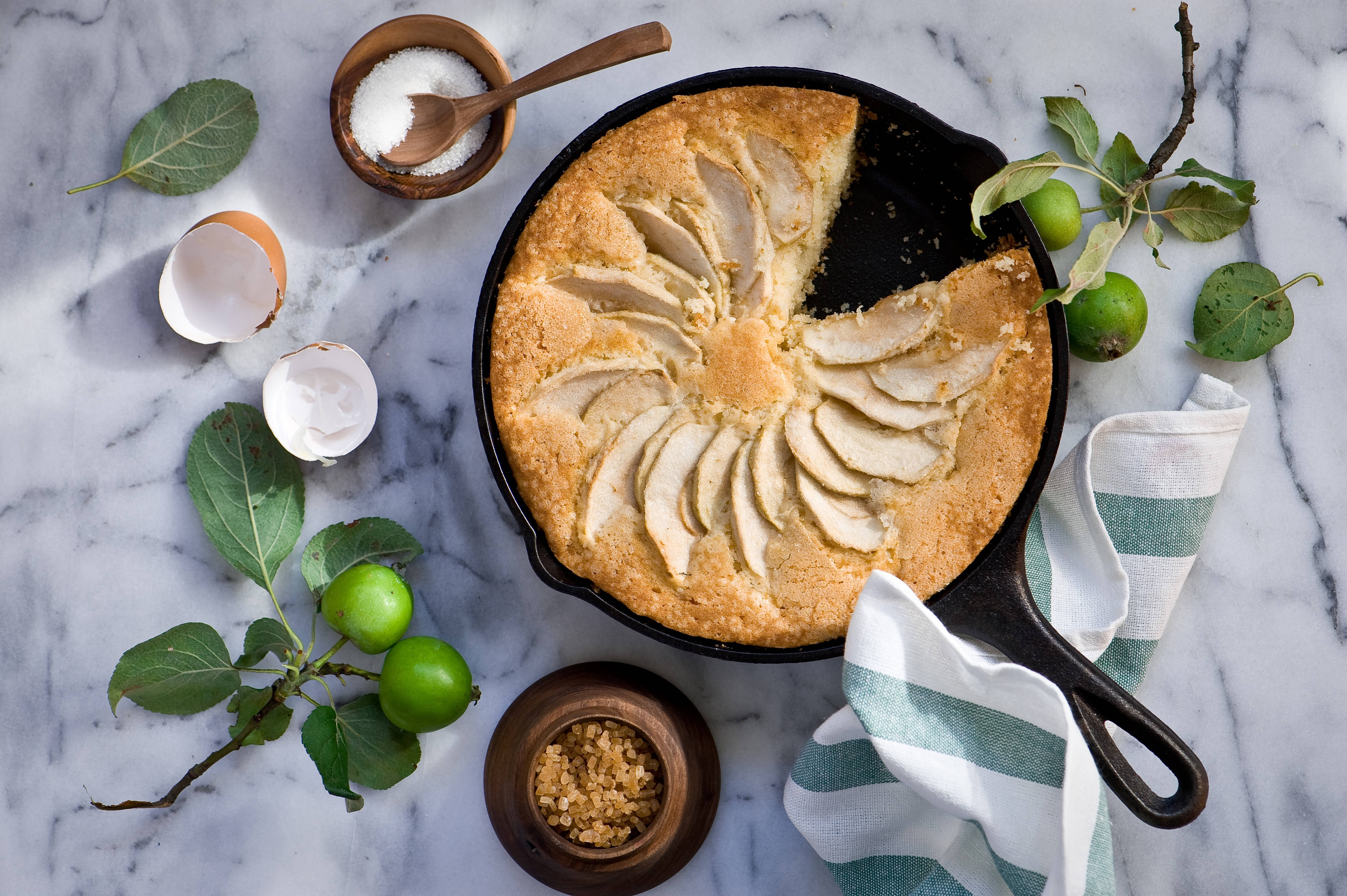 Pie 桌布 壁紙 — 一個金黃色的蘋果蛋糕放在鑄鐵煎鍋中，置於大理石表面上，周圍環繞著新鮮的青蘋果、樹葉和蛋殼。