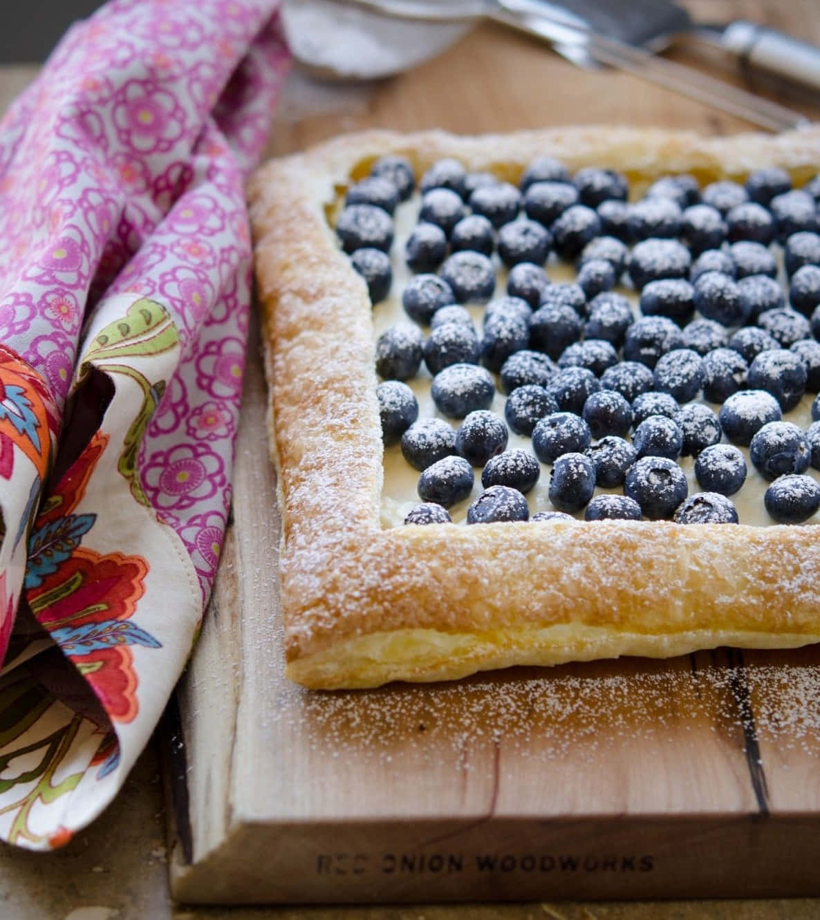 Blueberries Tart 桌布 壁紙 — 一個金黃色的酥皮塔，上面鋪滿新鮮藍莓和糖粉，放在木板上，旁邊有一塊色彩繽紛的花布。