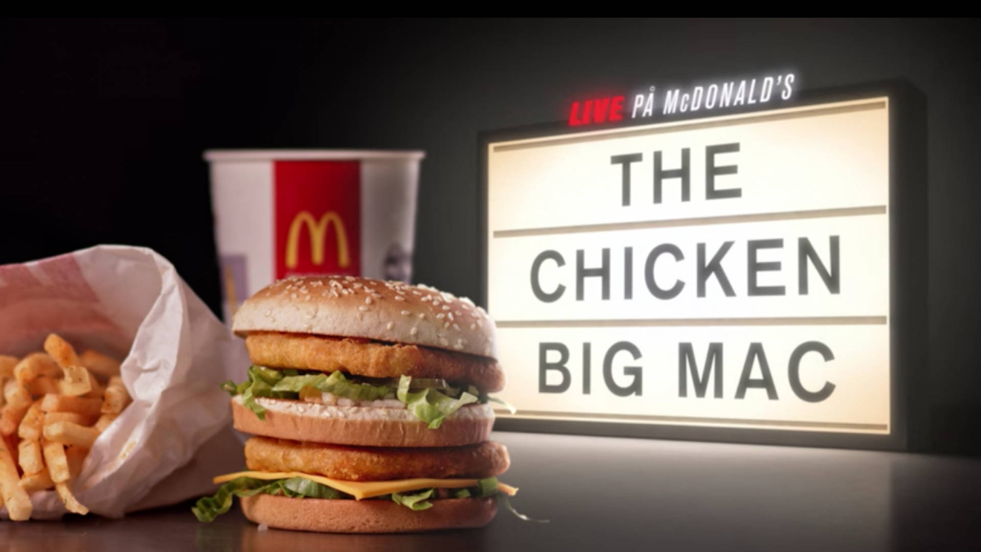 Mcdonalds 桌布 壁紙 — 一個金黃色的雞肉大麥克漢堡，搭配薯條和飲料杯，放在一個黑暗的攝影棚環境中，前面有一個發光的招牌顯示其名稱。