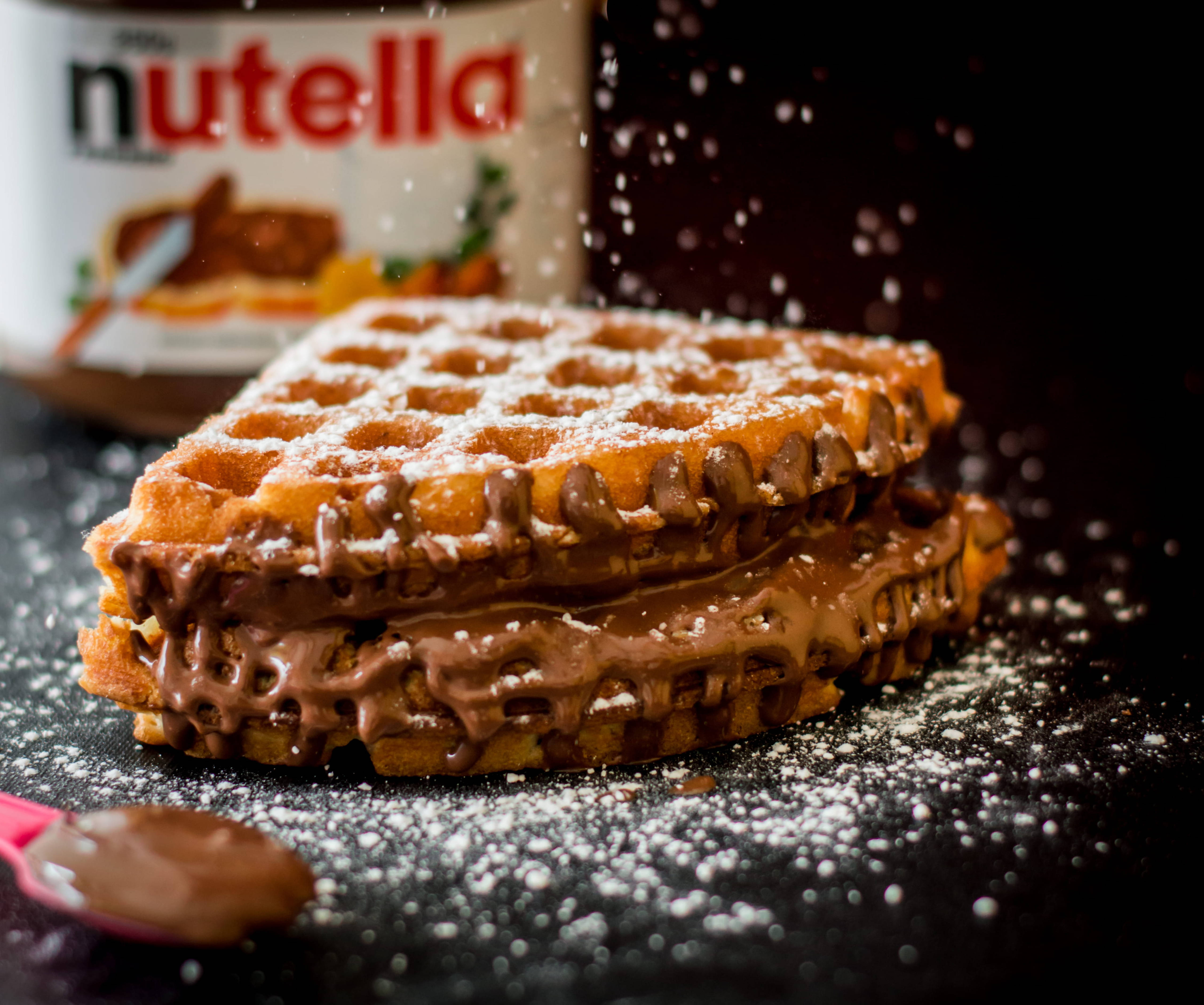 Waffle 桌布 壁紙 — 一個金黃色的鬆餅三明治，流出Nutella，糖粉在深色背景前於空中飄落。