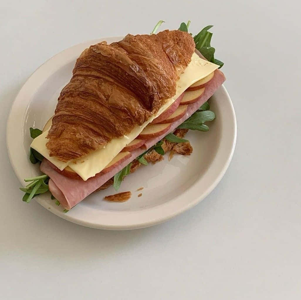 Minimalist Food 桌布 壁紙 — 一個金黃酥脆的可頌三明治，夾著火腿、起司和芝麻葉，放在白色盤子上，背景簡約。