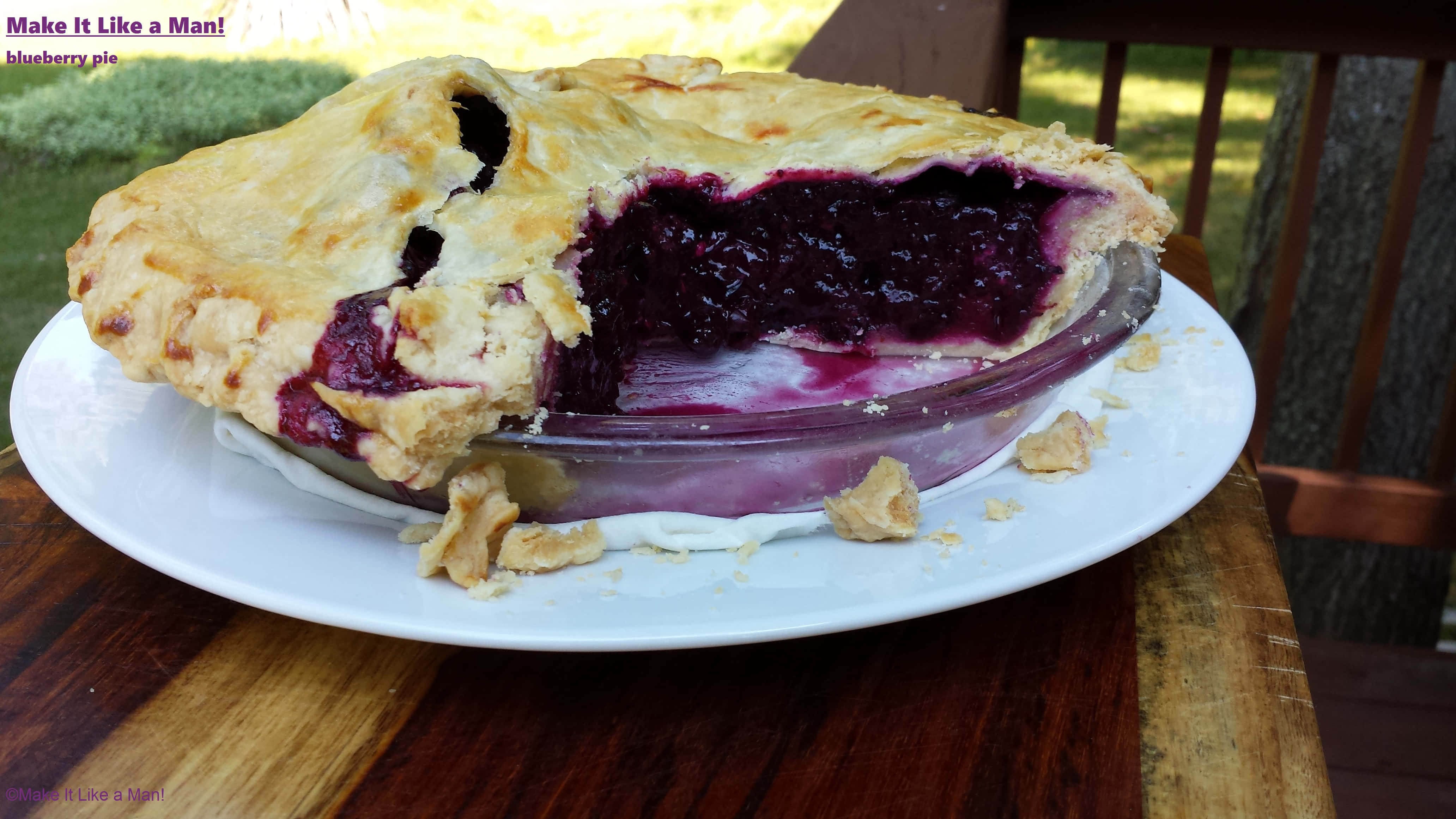 Blueberry Pie 桌布 壁紙 — 一個金黃酥脆的藍莓派，被切走了一塊，放在白色盤子上，置於戶外木桌上。
