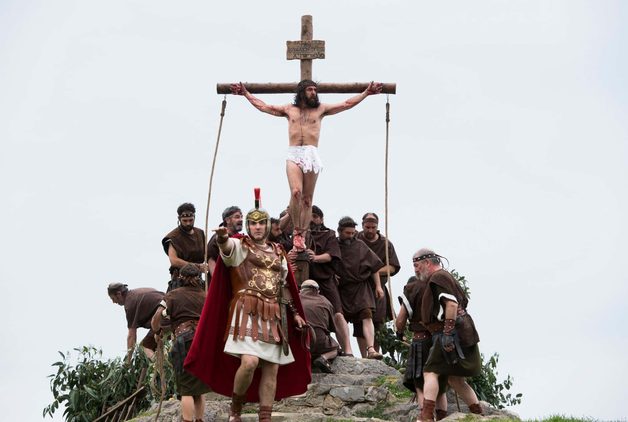 Good Friday 桌布 壁紙 — 一個釘十字架的場景，耶穌在木十字架上，被羅馬士兵和旁觀者包圍，在岩石山丘上，天空多雲。