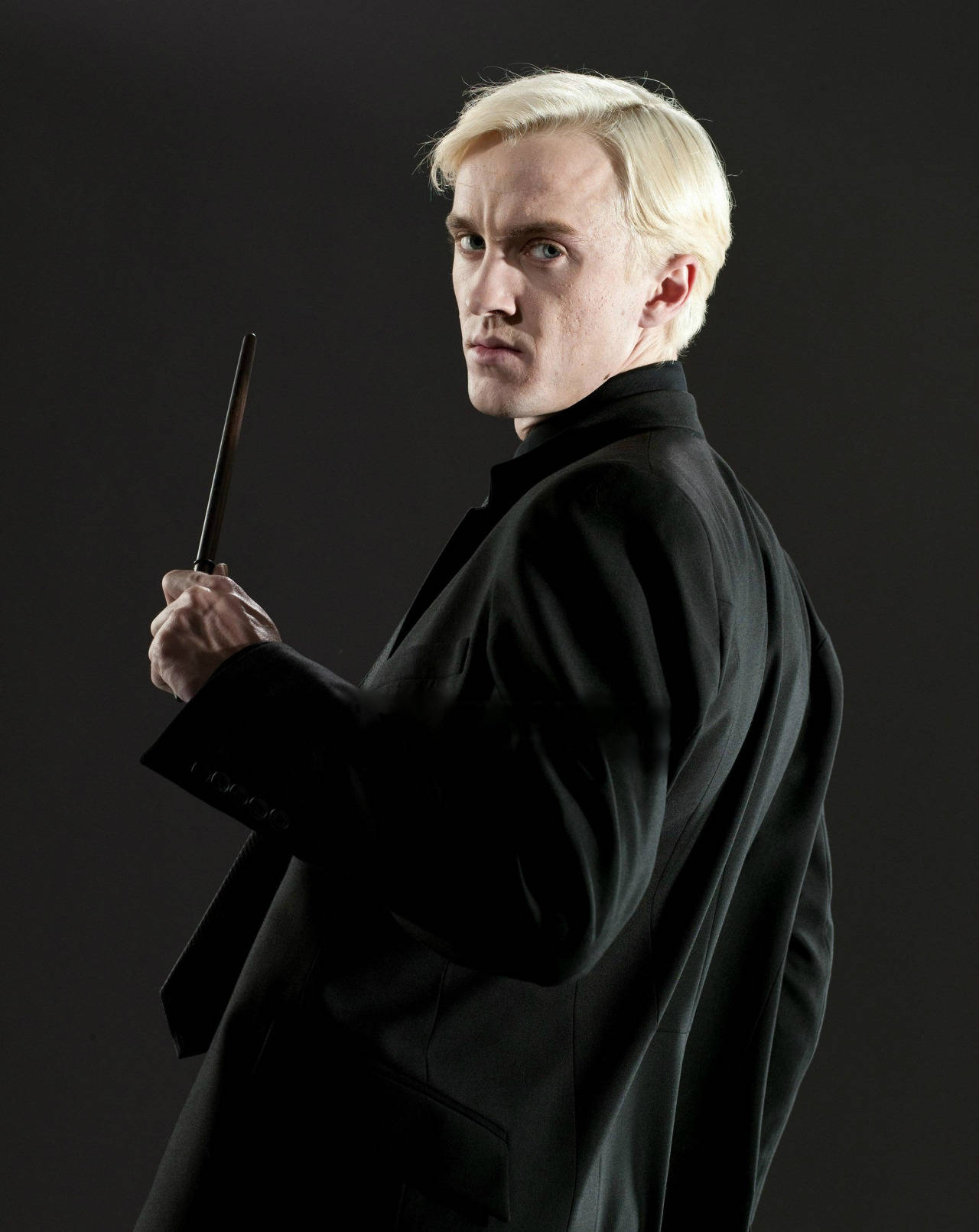 draco 桌布 壁紙 — 一個鉑金色頭髮的男人穿著黑色長袍，手持魔杖，在黑暗背景下回頭望向肩膀後方。